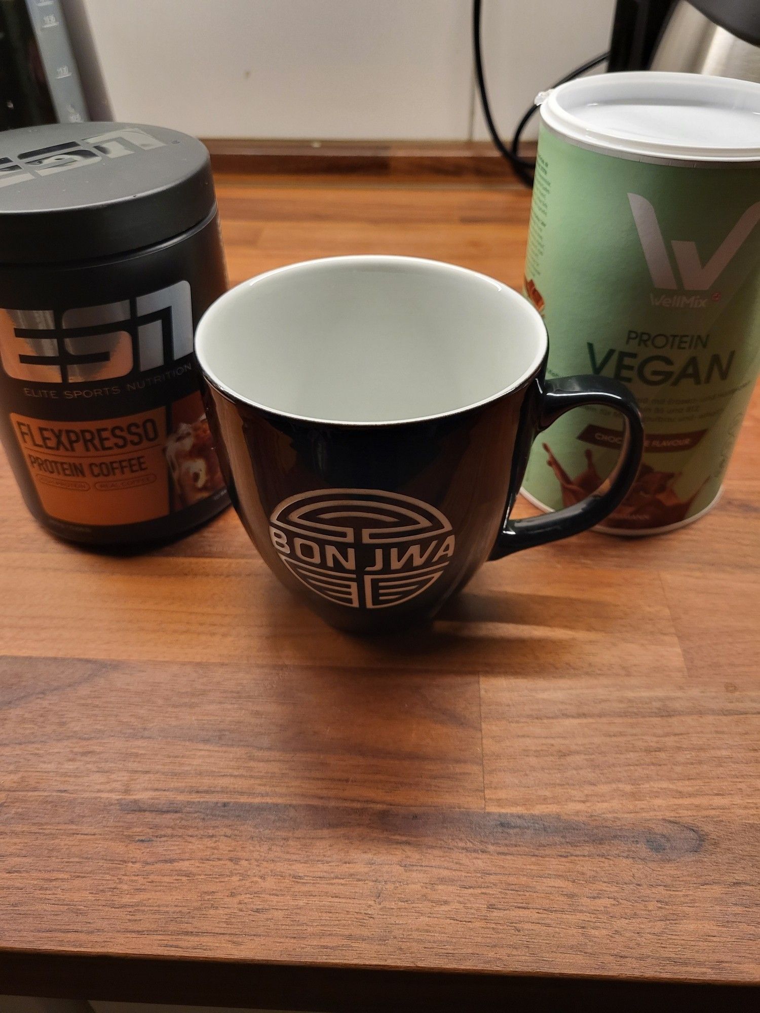 Man sieht die Arbeitsplatte einer Küche, darauf stehen eine schwarze Tasse mit weißem "Bonjwa" Aufdruck. Daneben eine Dose ESN Protein Kaffee und eine grüne Dose Vegan Protein