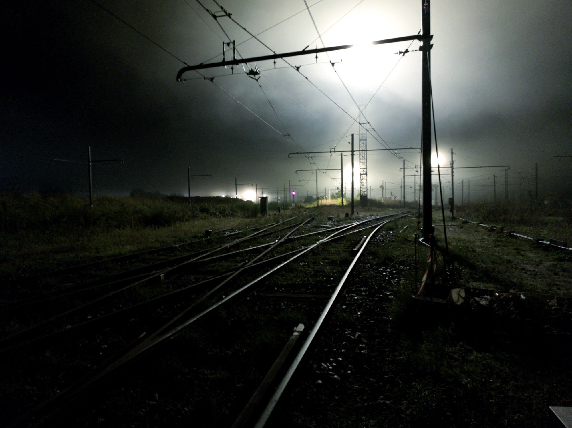 Photo de mes voies de train à mon arrivée aux alentours de 4h du matin. C'est très nuageux, il y'a un épais brouillard et on voit au loin des spots lumineux brisant la noirceur de la nuit et donnant un aspect pâle et fantomatique à la scène. Globalement, c'est une ambiance assez glauque du genre un clown tenant un ballon dans une bouche d'égout.