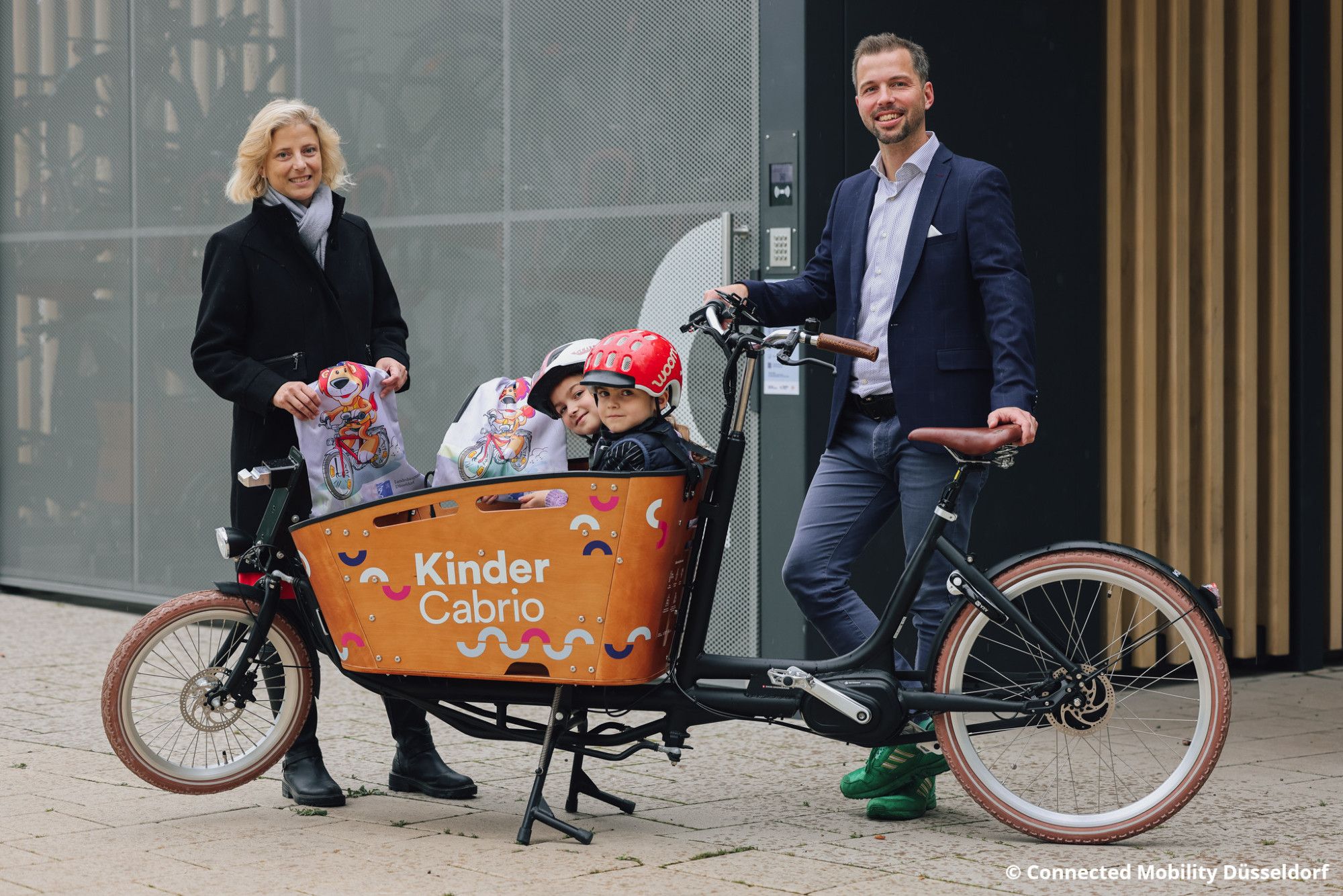 Katharina Metzker, Leiterin des Amtes für Verkehrsmanagement, steht gemeinsam mit Dr. David Rüdiger, Geschäftsführer der Connected Mobility, hinter einem Lastenrad. In dessen Korb, der mit der Aufschrift "Kinder Cabrio" versehen ist, sitzen zwei Kinder mit Helmen.