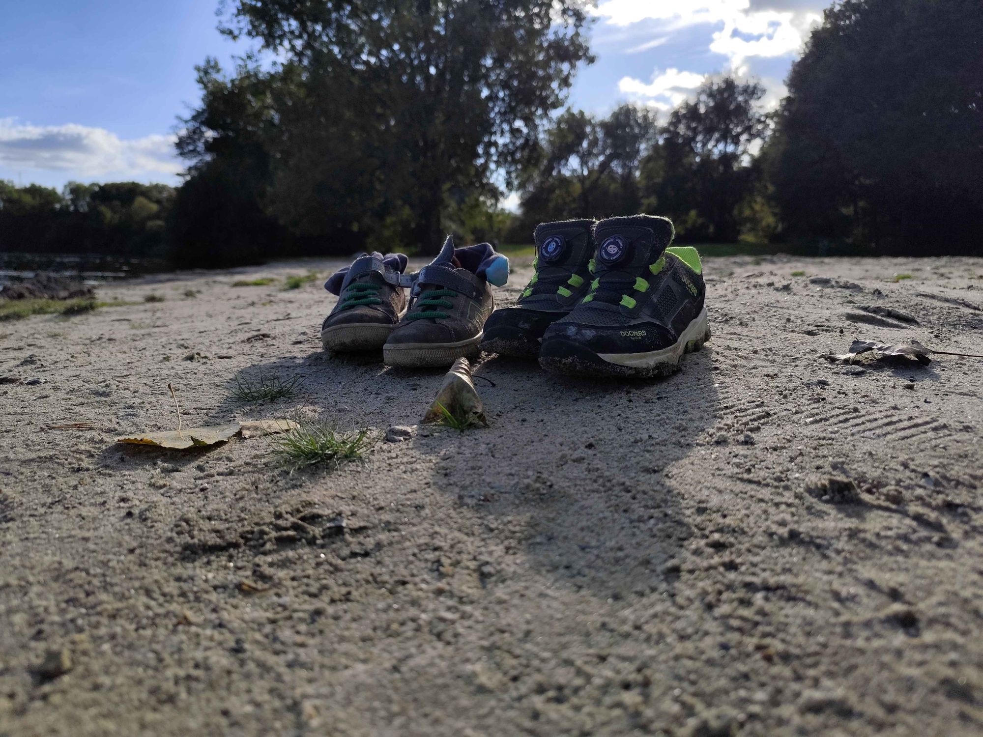 Zwei Paar Kinderschuhe im Sand aus der Froschperspektive. Im Hintergrund Büsche und Bäume und blauer Himmel mit ein paar Wolken.