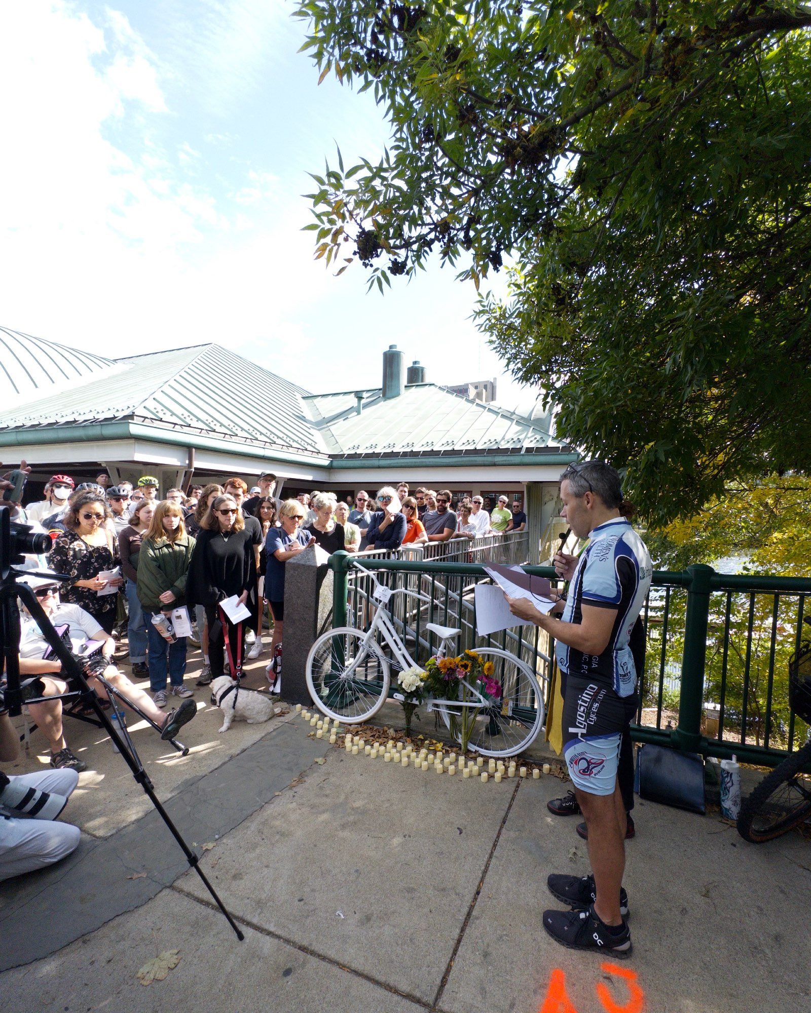 A photograph of a crowd of people standing around a white-painted ghost bicycle surrounded by candles, faced by a spandex-clad man speaking about his friend John Corcoran, who had been killed days before on the same spot.