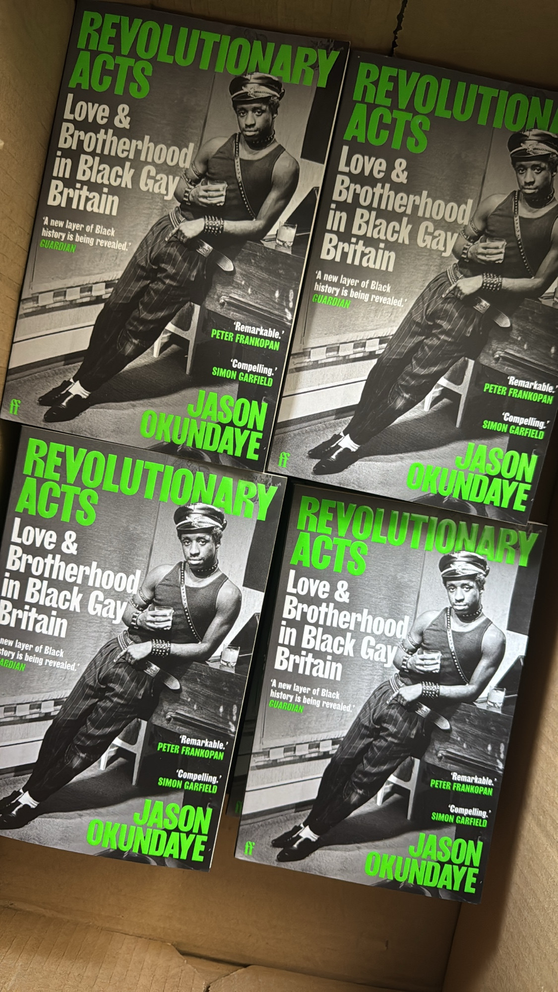 Paperback copies of Jason Okundaye’s book Revolutionary Acts. The cover features the title and the authors name in green writing and it is overload with a black and white image of a black man wearing a tank top, trousers and studded leather gear.