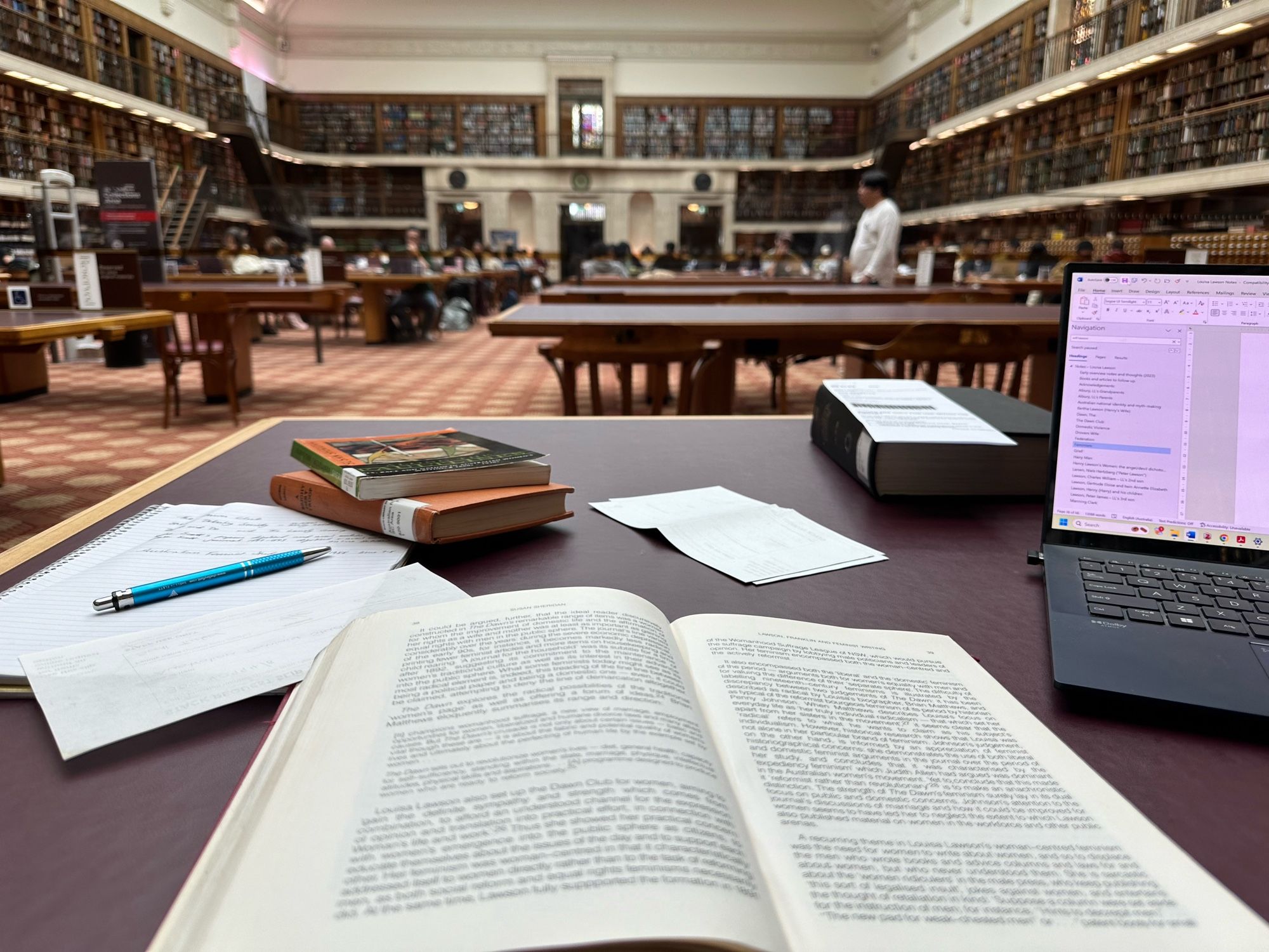 My photo of the Mitchell Reading Room at the State Library of NSW. My desk in the foreground with open books and laptop, a large and ornate book-lined room in the background, filled with people studying.