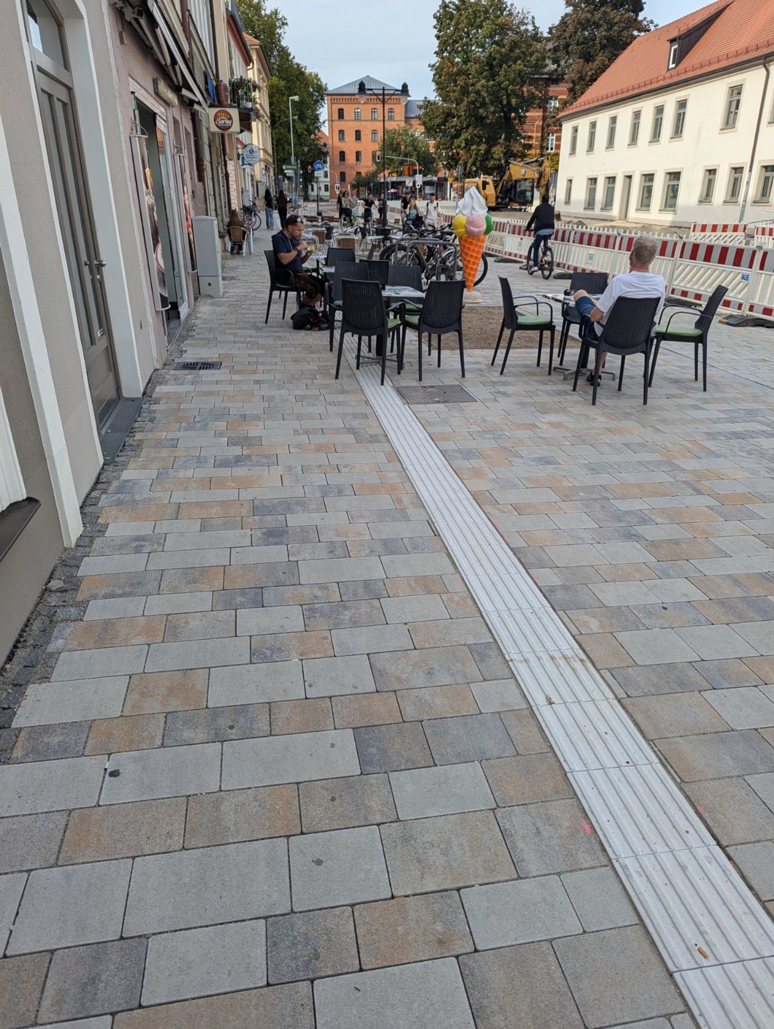 A wide, newly done sidewalk with a white tactile marker for guiding visually impaired, leading directly into a group of chairs for outside seating of an ice cream parlor.