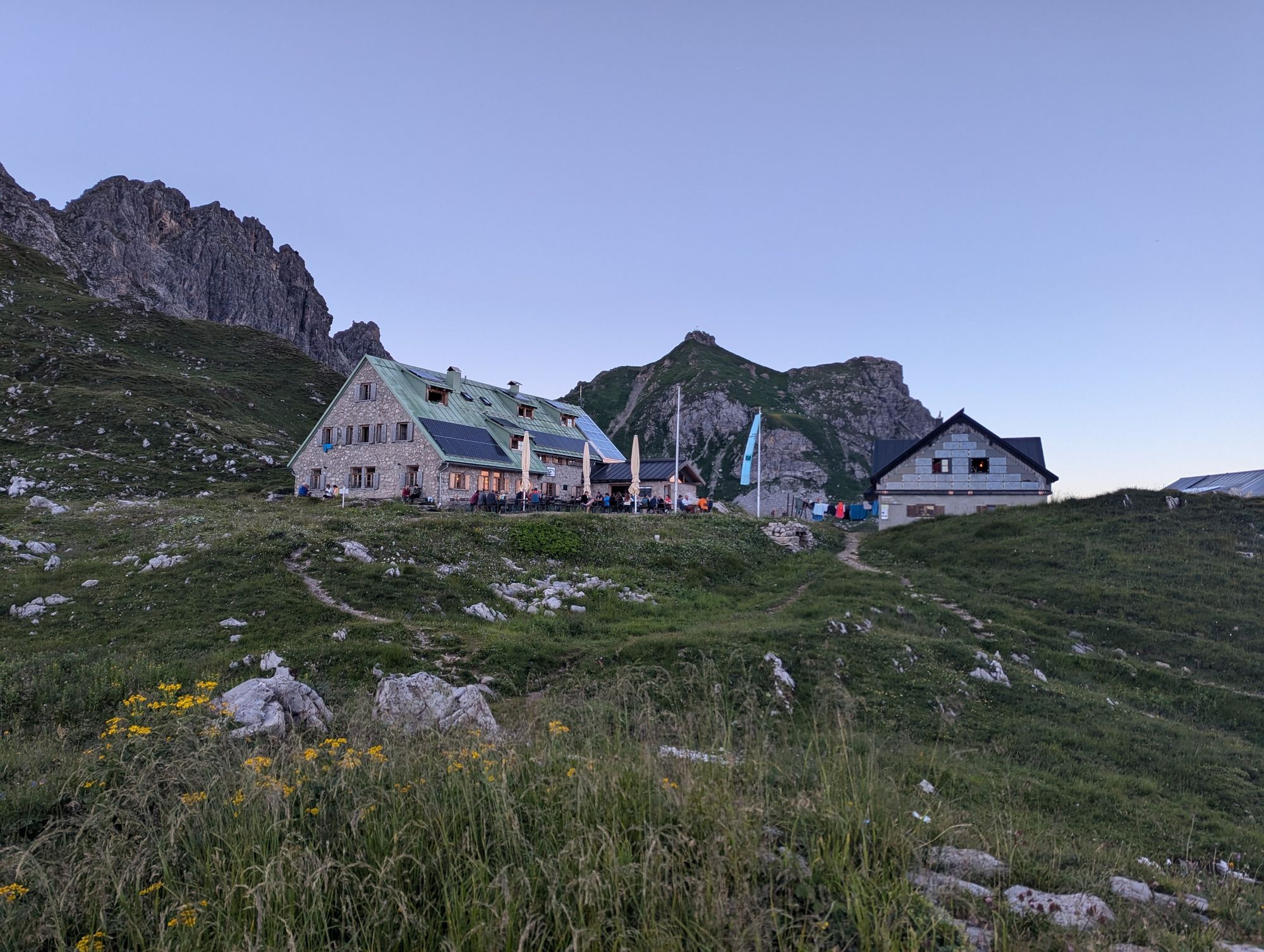 An assembly of two huts, the smaller one has a a big assembly of solar panels on its side, while on the bigger one the solar panels contrast with the green copper roof. The foreground is all green with patches of limestone and behind the hut bigger limestone rocks reach into the sky.
The whole picture is tinted violet from the late evening sun