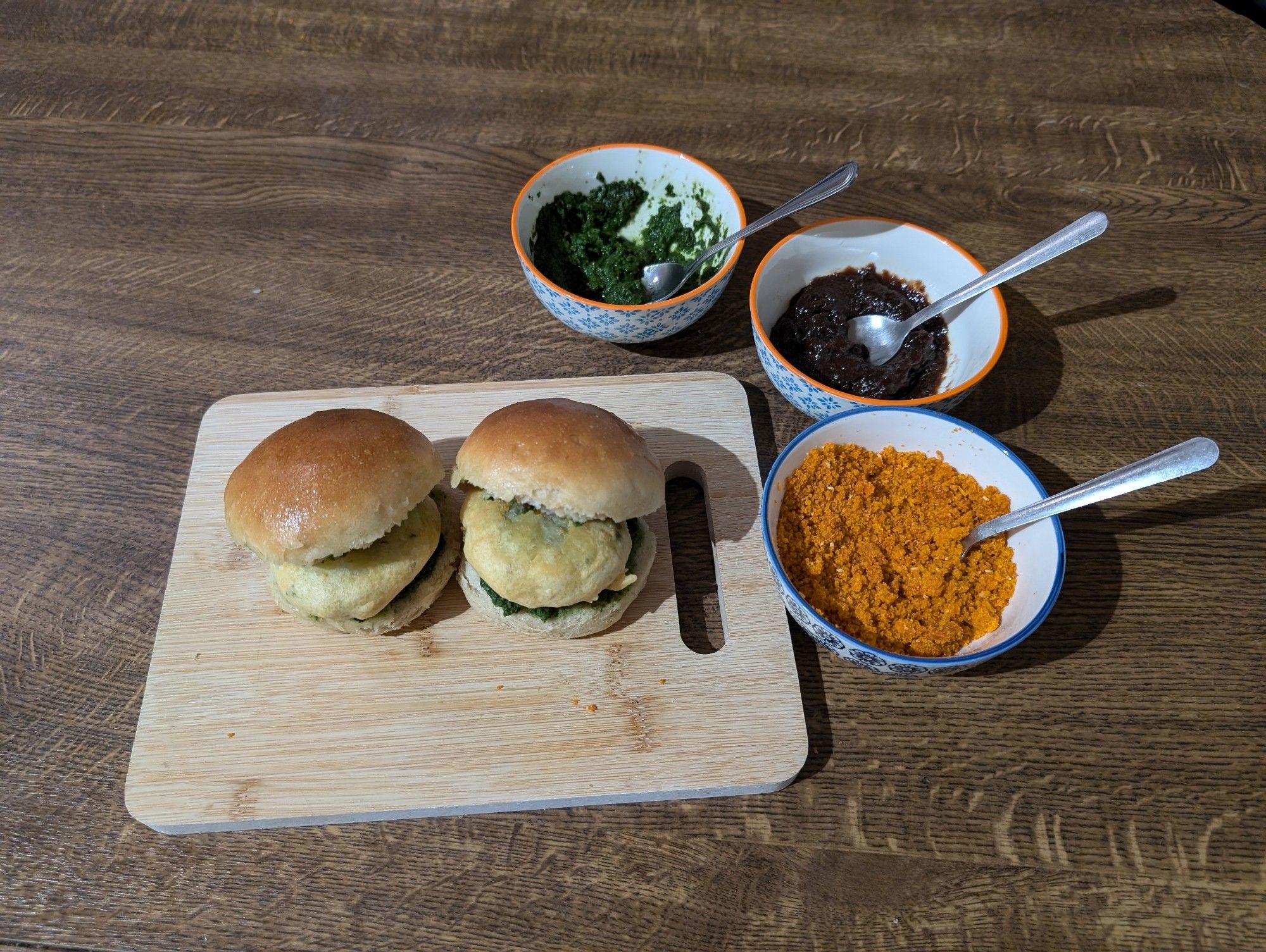 Photo of two Vada Pavs on a cutting board. These are sandwiches made of soft bread rolls, filled with deep-fried battered mashed potato and several chutneys. Small bowls of the chutneys I made and used are next to the board. These are a dark green spicy coriander chutney, a dark brown tamarind chutney and a dry garlic chutney.