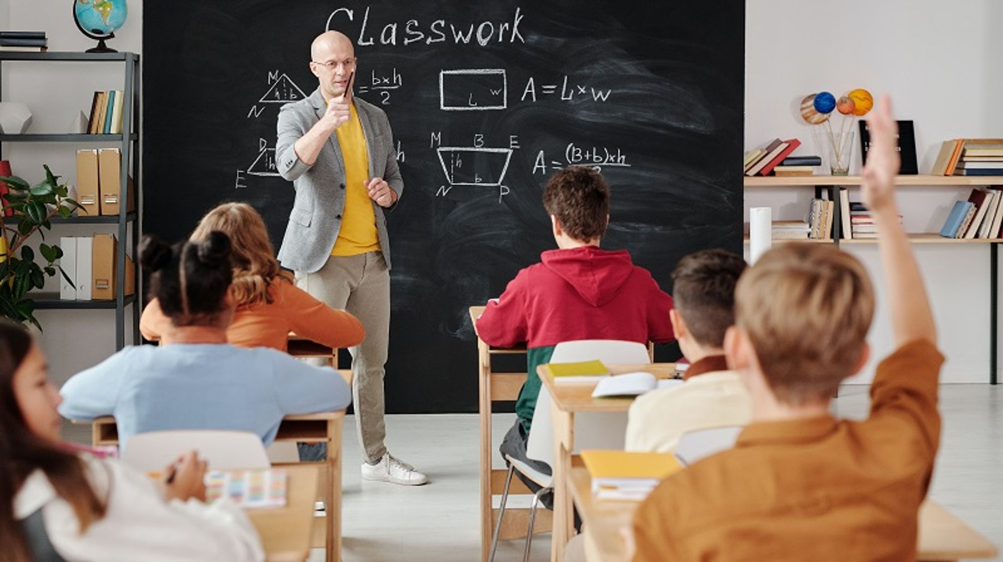 Bildbeschreibung: Ein Klassenzimmer mit Schüler/innen und vorne steht ein Lehrer an der Tafel.