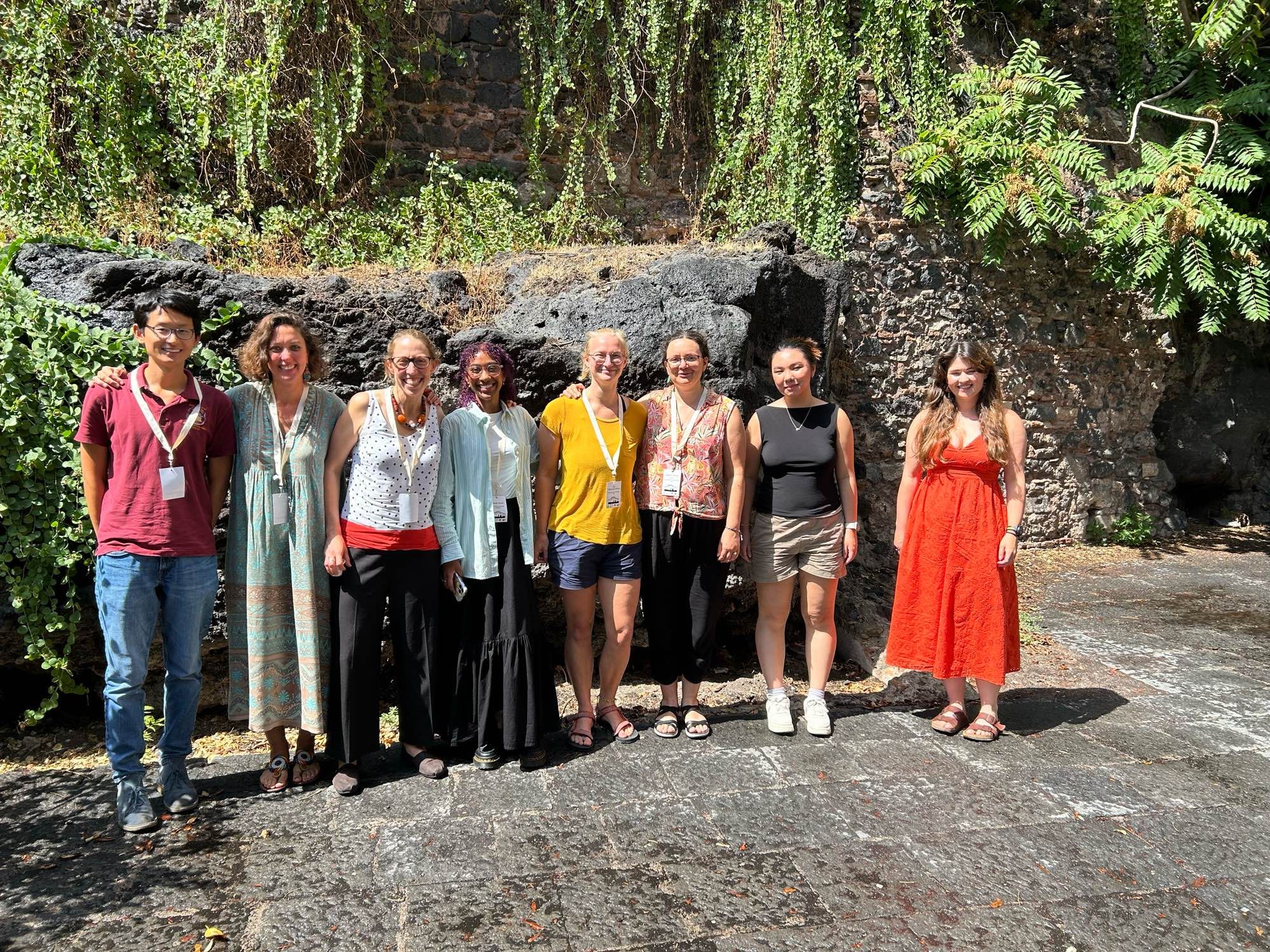 8 humans stood smiling in front of a wall built into a lava flow.