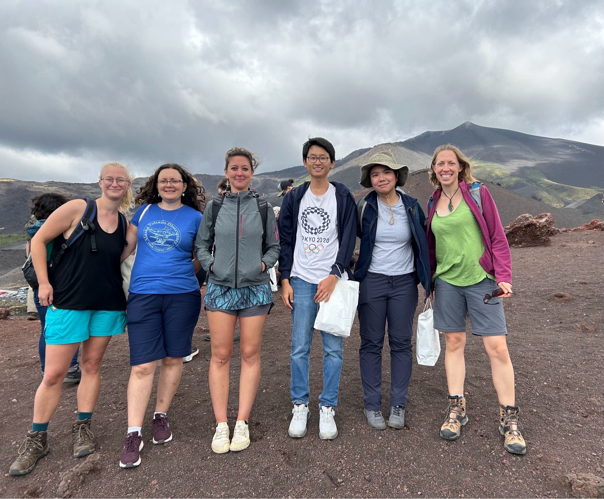 6 people in outdoor gear stood on ashy ground with a mountain/volcano in the background. It’s cloudy, but clear from shorts and sone bare arms that it is still quite warm.