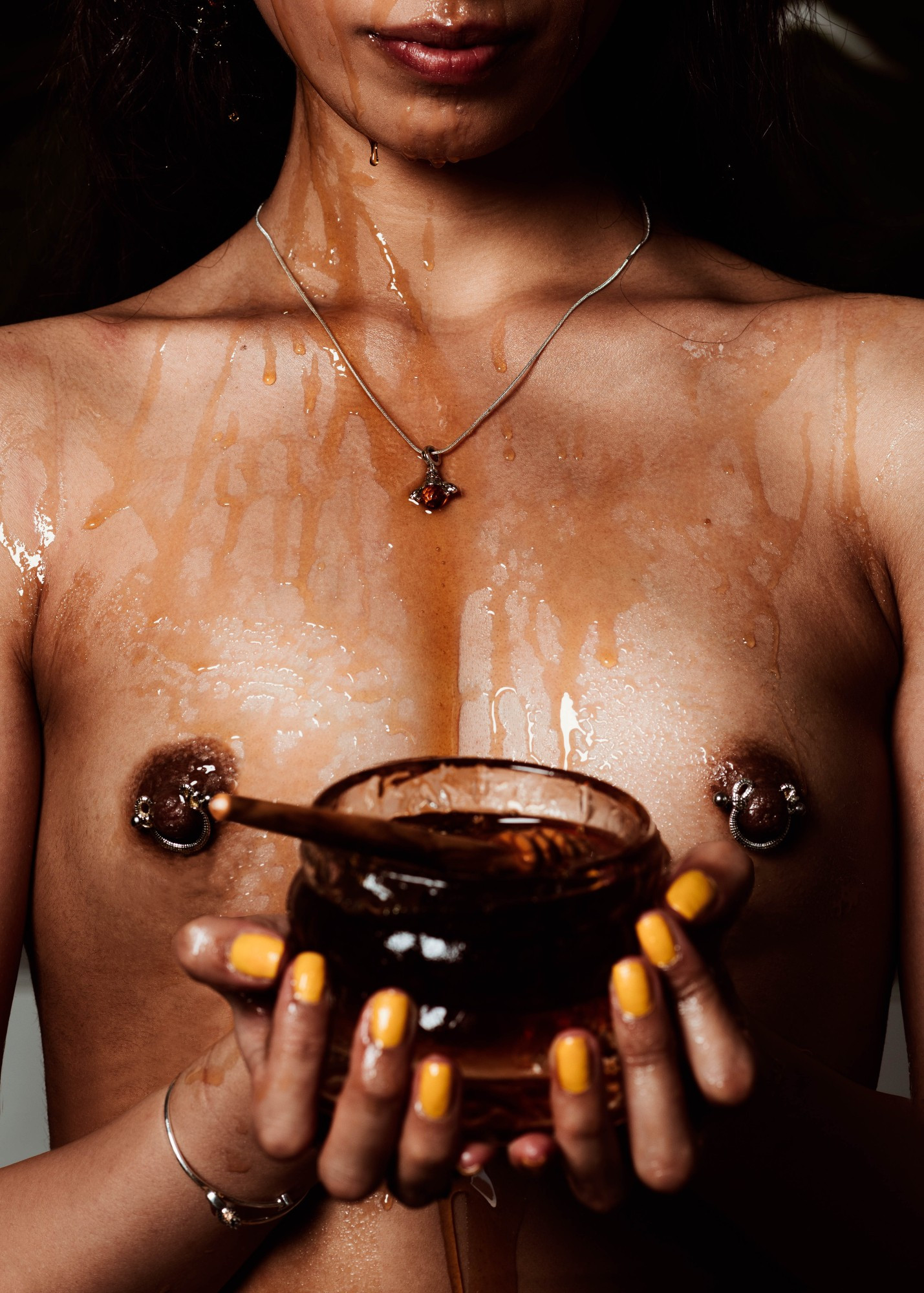 Model Nicole, pictured midriff up, holding a container filled with honey at chest height. Topless, nipples exposed and pierced. Upper body covered in honey, wearing a bee necklace. Cropped only showing lips