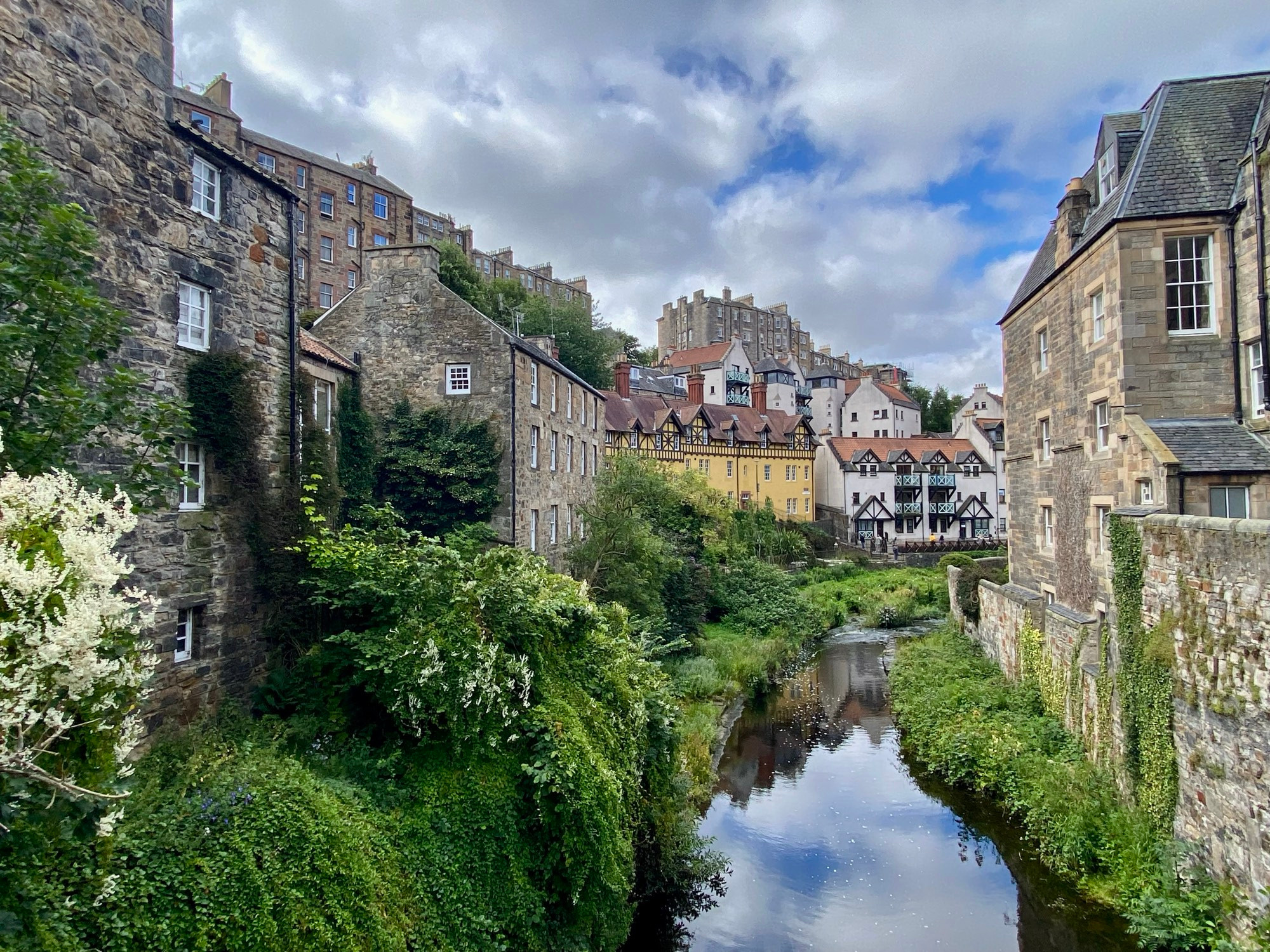 A picturesque view of Dean Village in Edinburgh, Scotland, showing historic stone buildings along a lush, green riverside with the Water of Leith flowing through the center. Colourful houses and a blue sky with scattered clouds.