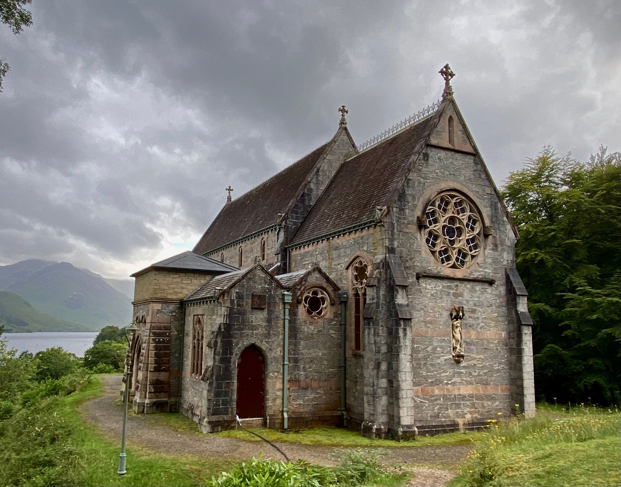 A stone Gothic-style church (The Catholic Church of St Mary and St Finnan) with stained glass windows and a steep slate roof, set against a backdrop of mountains and Loch Shiel in Glenfinnan, Scotland.