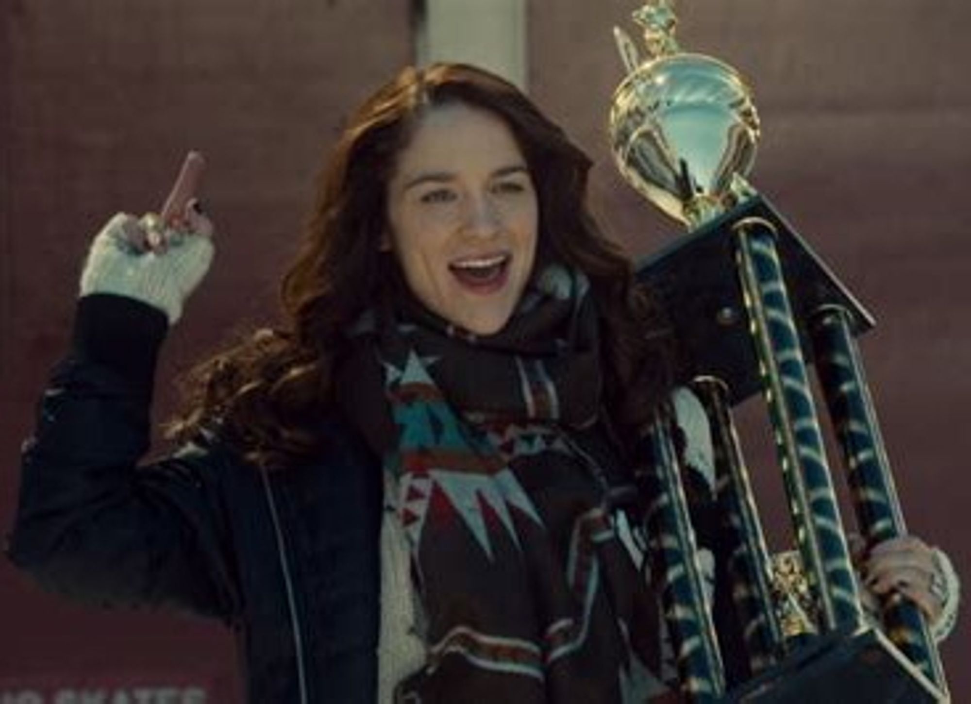 An image of Melanie Scrofano as Wynonna Earp. She is carrying a trophy in on hand while flipping the bird with the other. She is smiling and happy.