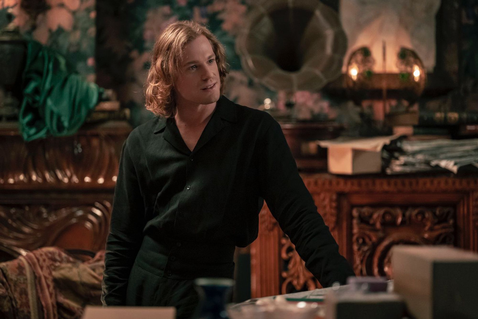 Still image of Lestat de Lioncourt from AMC's Interview with the Vampire, dressed in a black outfit and leaning against a table.