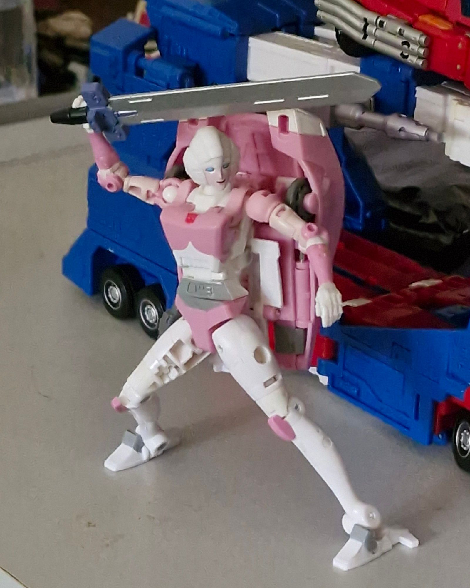 A photograph of a Transformers the Movie Studio Series Arcee toy, holding one of Springer's swords above her head, posed in a ready-for-battle stance.