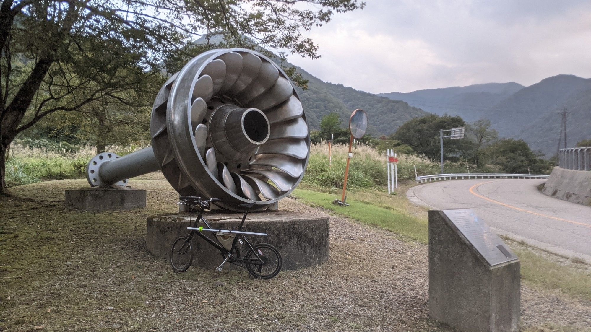 A defunct hydroelectric dam turbine on a plinth as a roadside attraction. There is a bicycle for scale, which is dwarfed by the turbine.