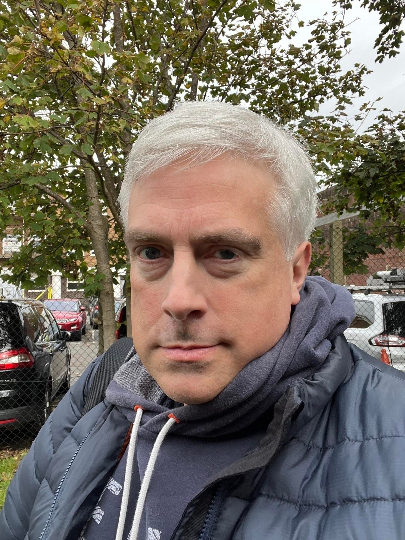 Adam Tinworth with neatly cut gray hair is outside in front of trees and parked cars, wearing a dark jacket and a hoodie.