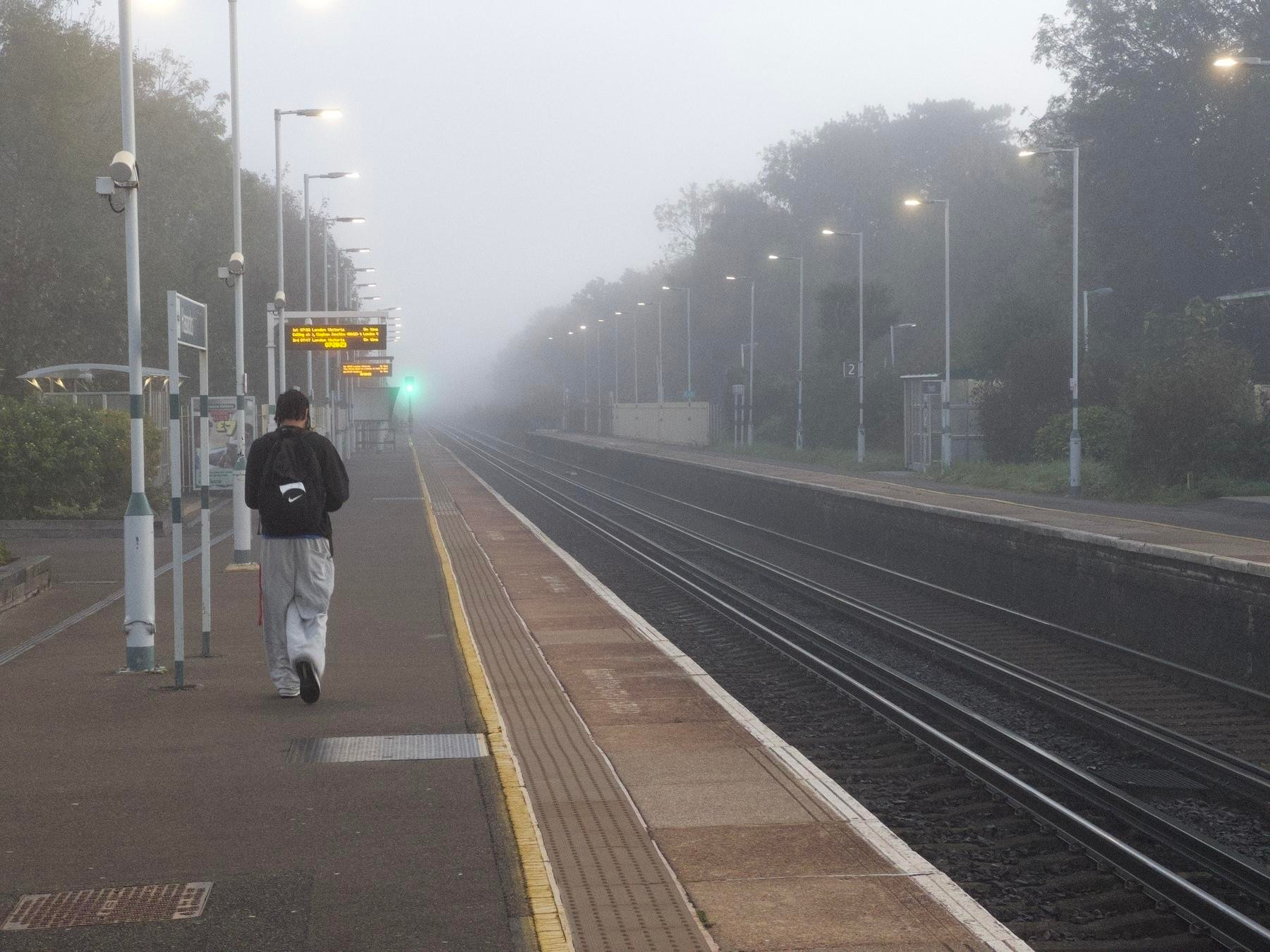 A person with a backpack walks along a foggy train platform, with tracks running parallel and a digital signboard in the distance.