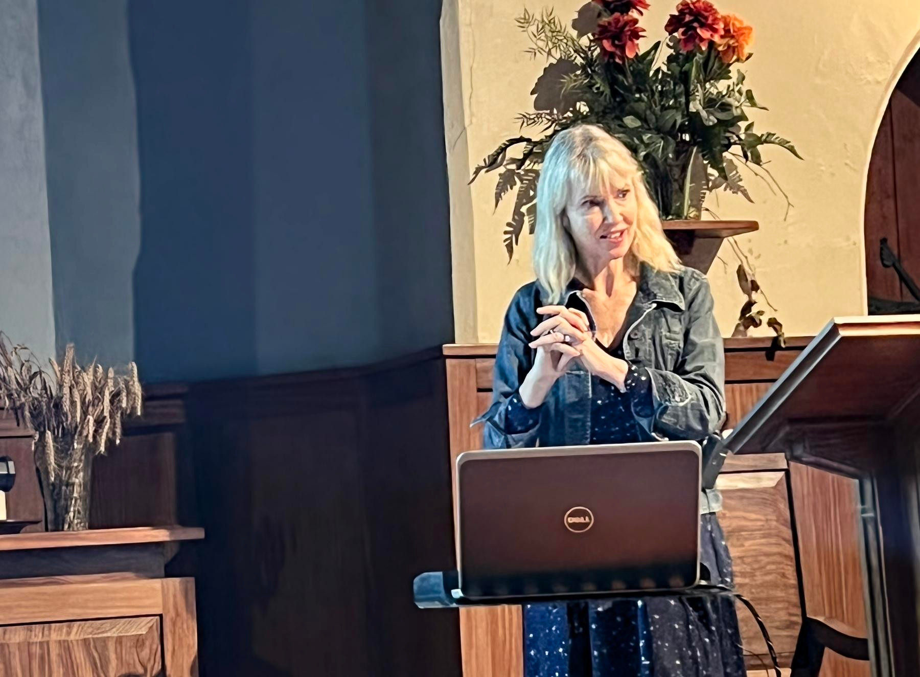 The author Annabel Abbs stands at a podium in Steyning Methodist Church, with a laptop, speaking in a room with wooden paneling and a floral arrangement in the background.