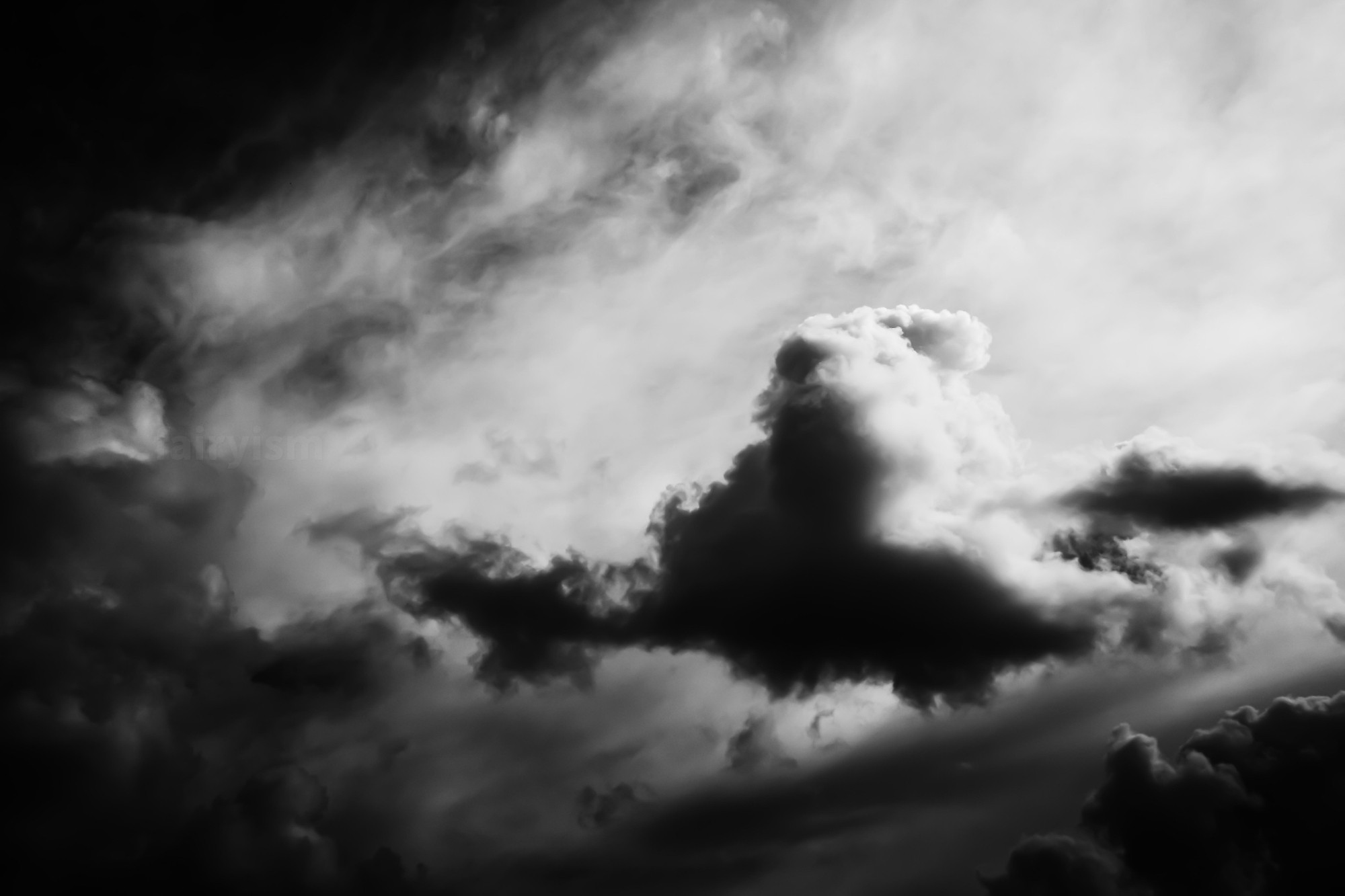 Dark black and white capture of the sky with clouds in it. One single puffy cloud is separated by the light in the middle of the image, leaning towards the left side.