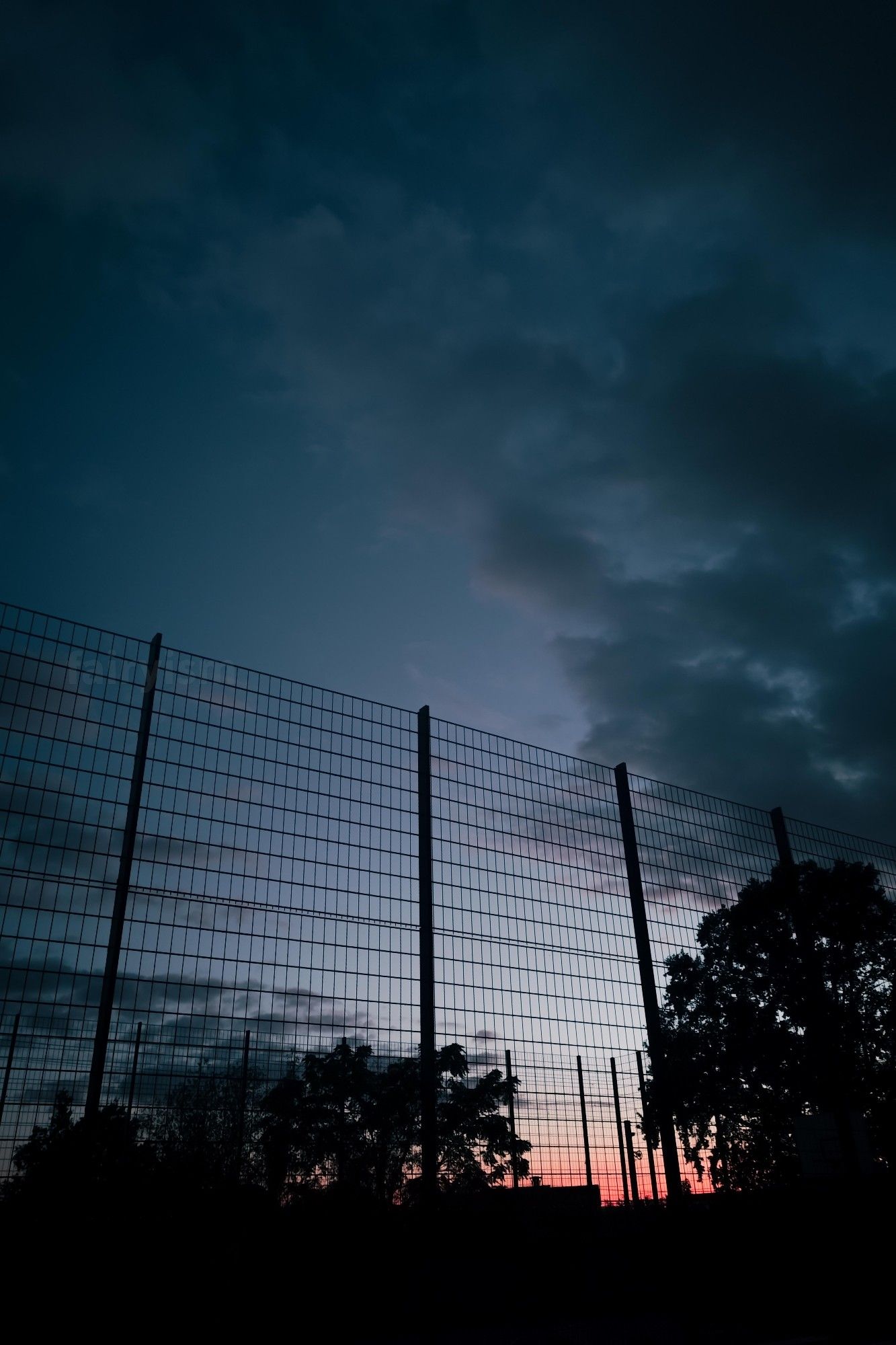 Vertical capture of a high fence, captured against the sunset sky during blue hour. In front of the fence are the silhouettes of trees and bushes, and in the sky are some clouds.