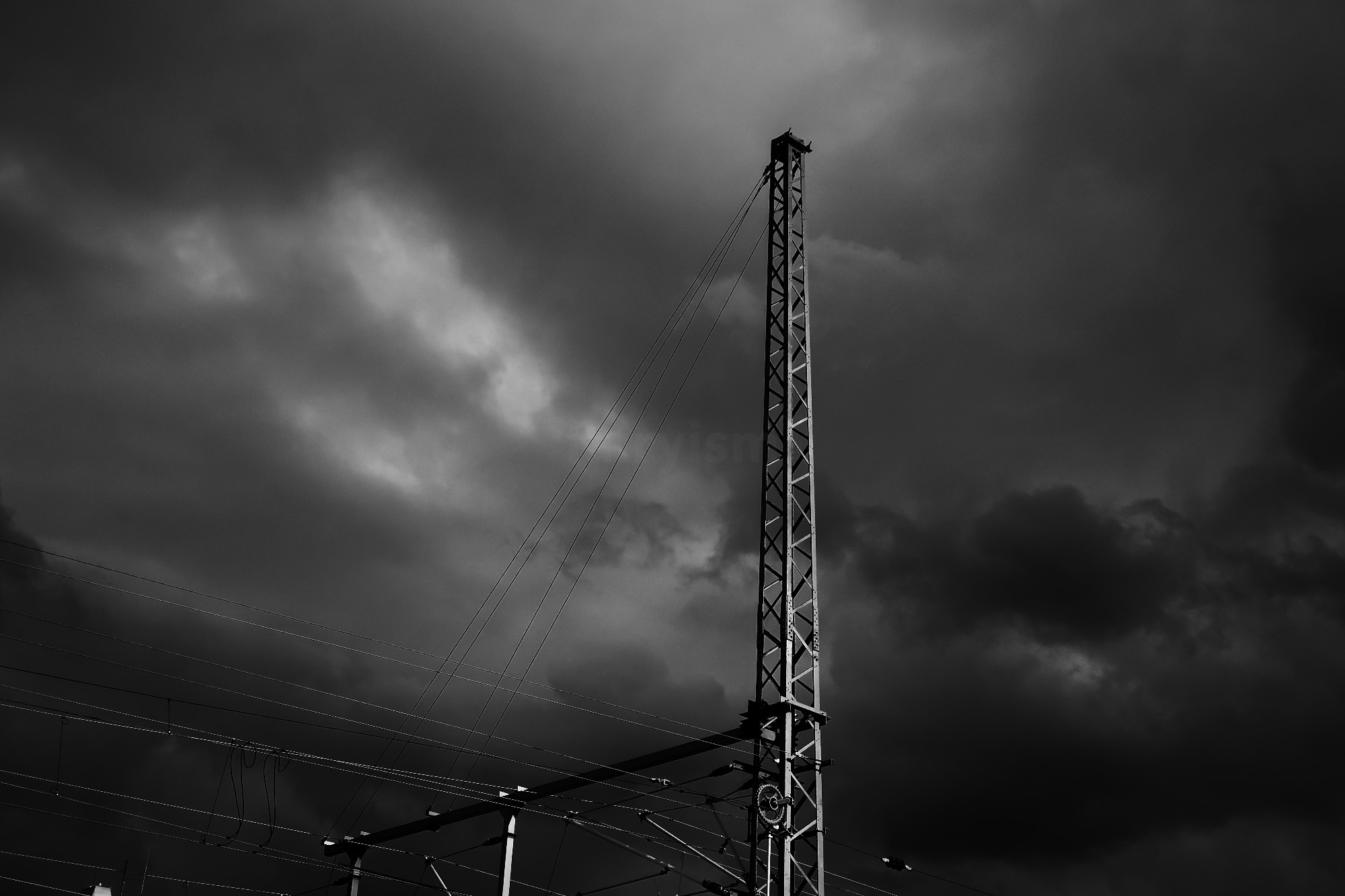 A power pole captured in black and white, with the cloudy sky in the background. At the bottom of the photo are also power lines.