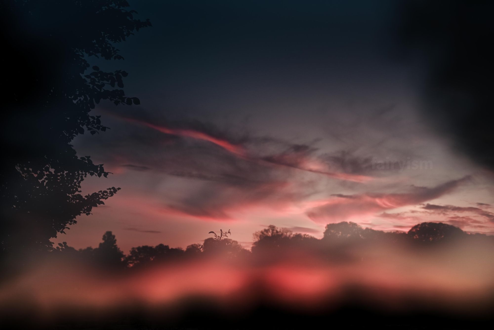 Photograph of the silhouettes of trees on the horizon, with the red and orange cloudy sunset sky above it. There is a blurry colourful sheen over the photo, which makes it appear a bit dreamy.