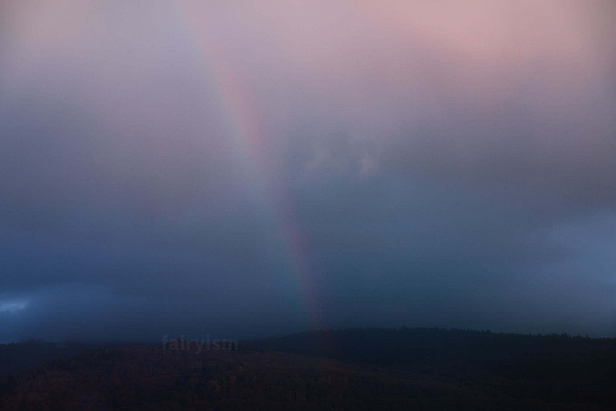 Capture of a small mountain range, with a soft rainbow pointing towards the middle of the horizon. The sky is very cloudy and covered, and the toning of the photo is a soft blue.