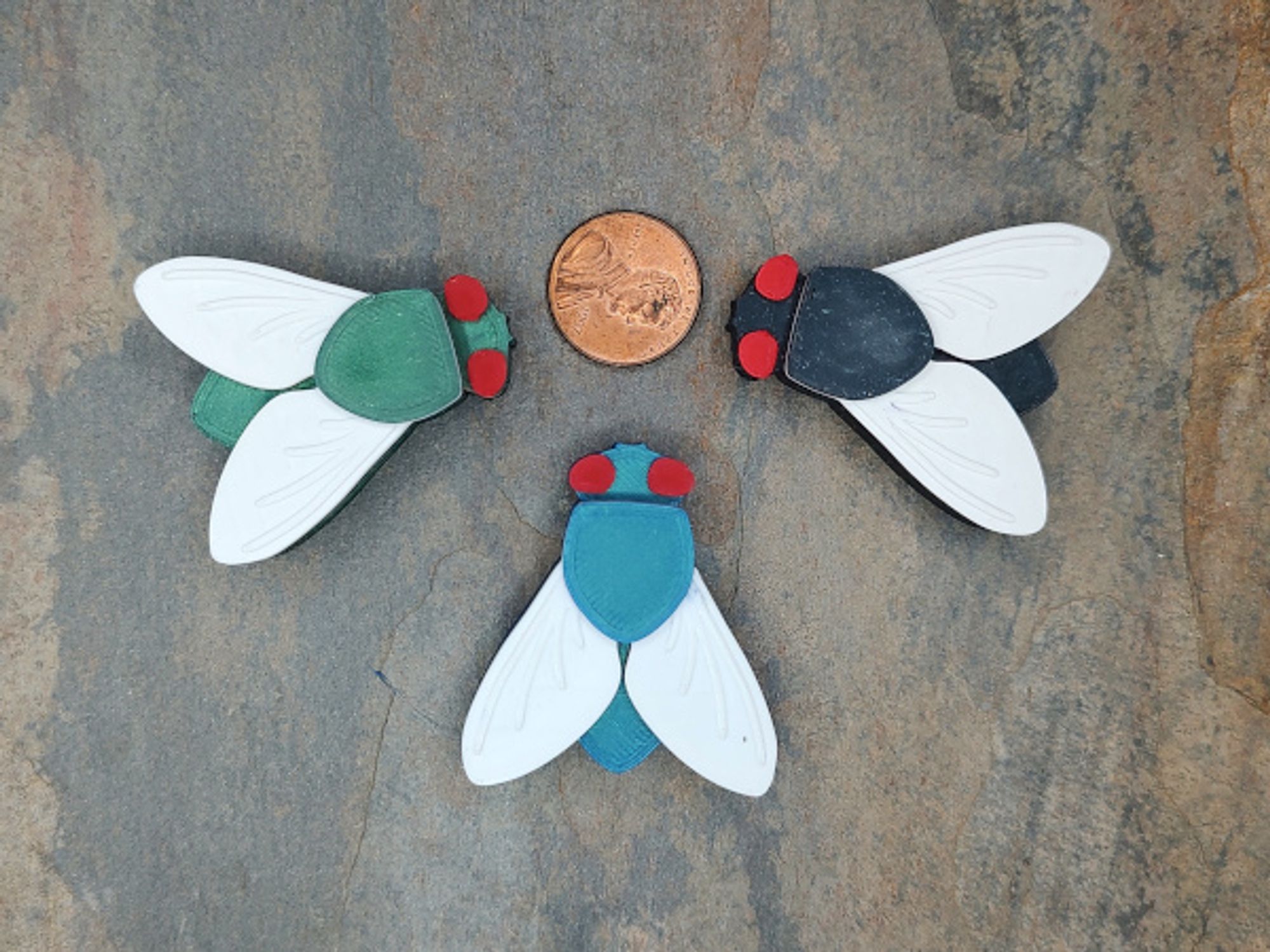 Three 3d printed stylized blowflies surrounding a US penny. They all have white wings and red eyes, but different colored bodies.  The left one is green, the bottom one is blue, and the right side is black.