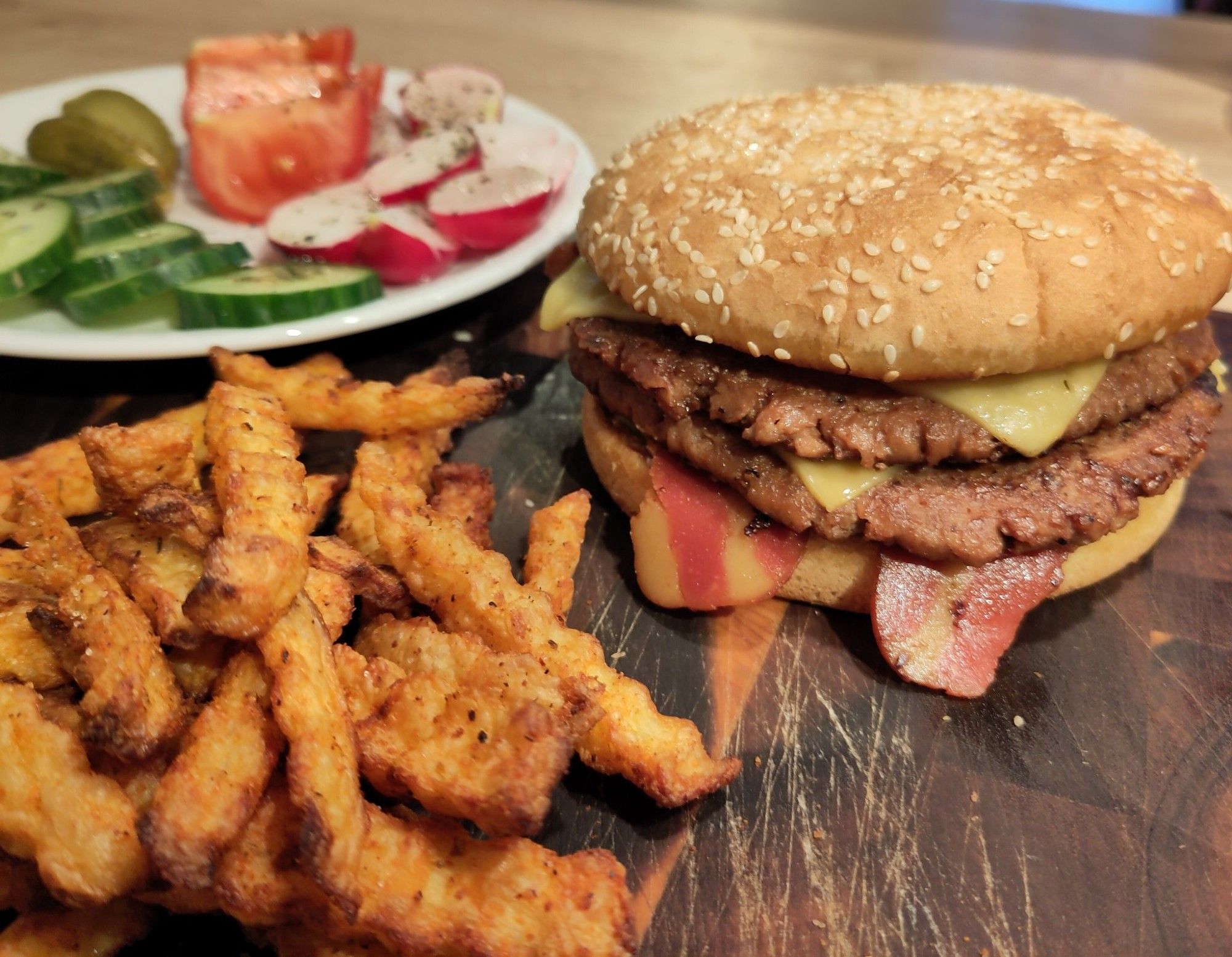 Rechts auf dunklem Schneidbrett ein Double Cheeseburger mit Bacon. Links im Vordergrund goldbraune Pommes und im Hintergrund ein Teller mit aufgeschnittenen Tomaten, Gurken, Pickels und Radieschen.