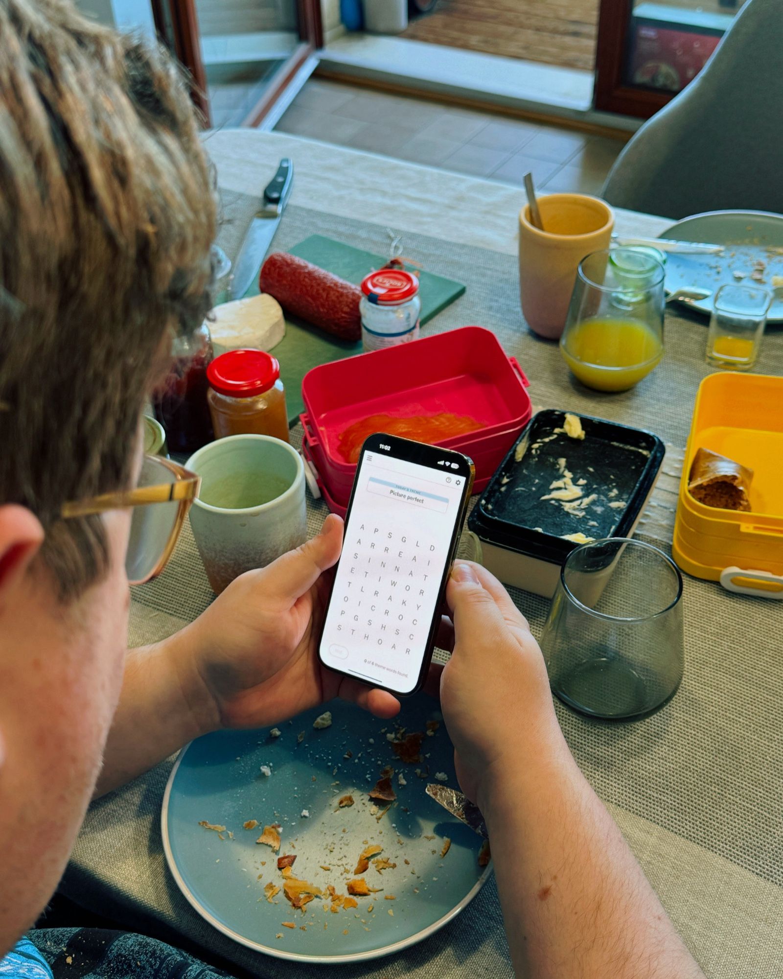 Über Christophers Schulter Richtung Frühstückstisch fotografiert – er hat ein Smartphone in der Hand, auf dem das NYT Rätsel „Strands“ zu sehen ist