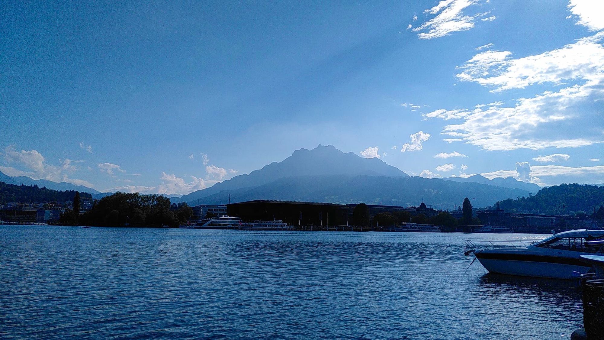 Seebild mit Berg im Hintergrund. Blauer Himmel mit ein paar weissen Wolken. Rechts ein Teil eines Bootes.