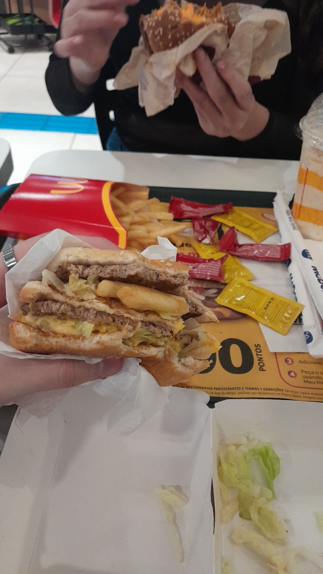 Duas pessoas saboreando seu próprio combo do McDonald's, com foco em um hambúrguer que tem batata frita como um dos ingredientes.