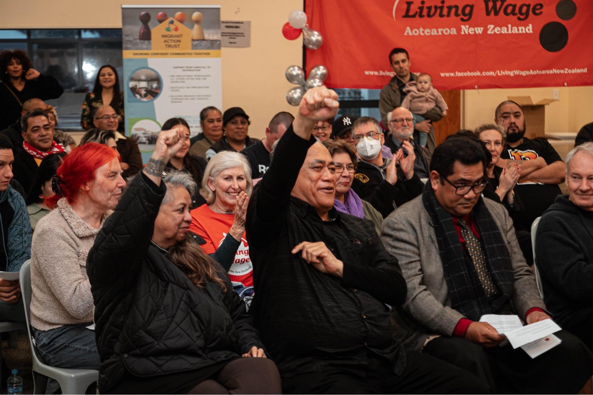 Seated smiling attendees at the Living Wage Election Forum in Auckland 2023. There’s a Living Wage and a Migrant Action Trust Banner in the background with red white and silver balloons. Next to the banners their are standing attendees.