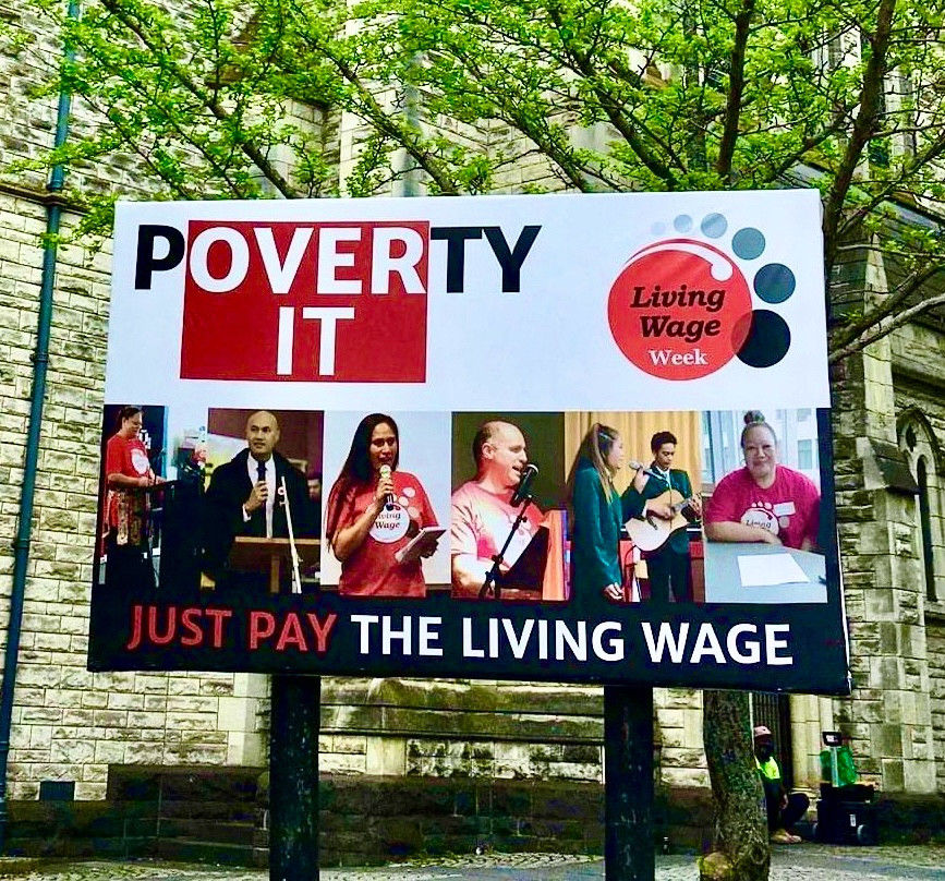 Billboard in front of a stone building with a tree directly behind the billboard. The centre of the billboards is a series of photos of speakers at Living Wage events, holding or in front of microphones. Above the photos the words. Poverty. Over It, to the left and to the right a large Living Wage Logo. Underneath the photo the words JUST PAY THE LIVING WAGE.