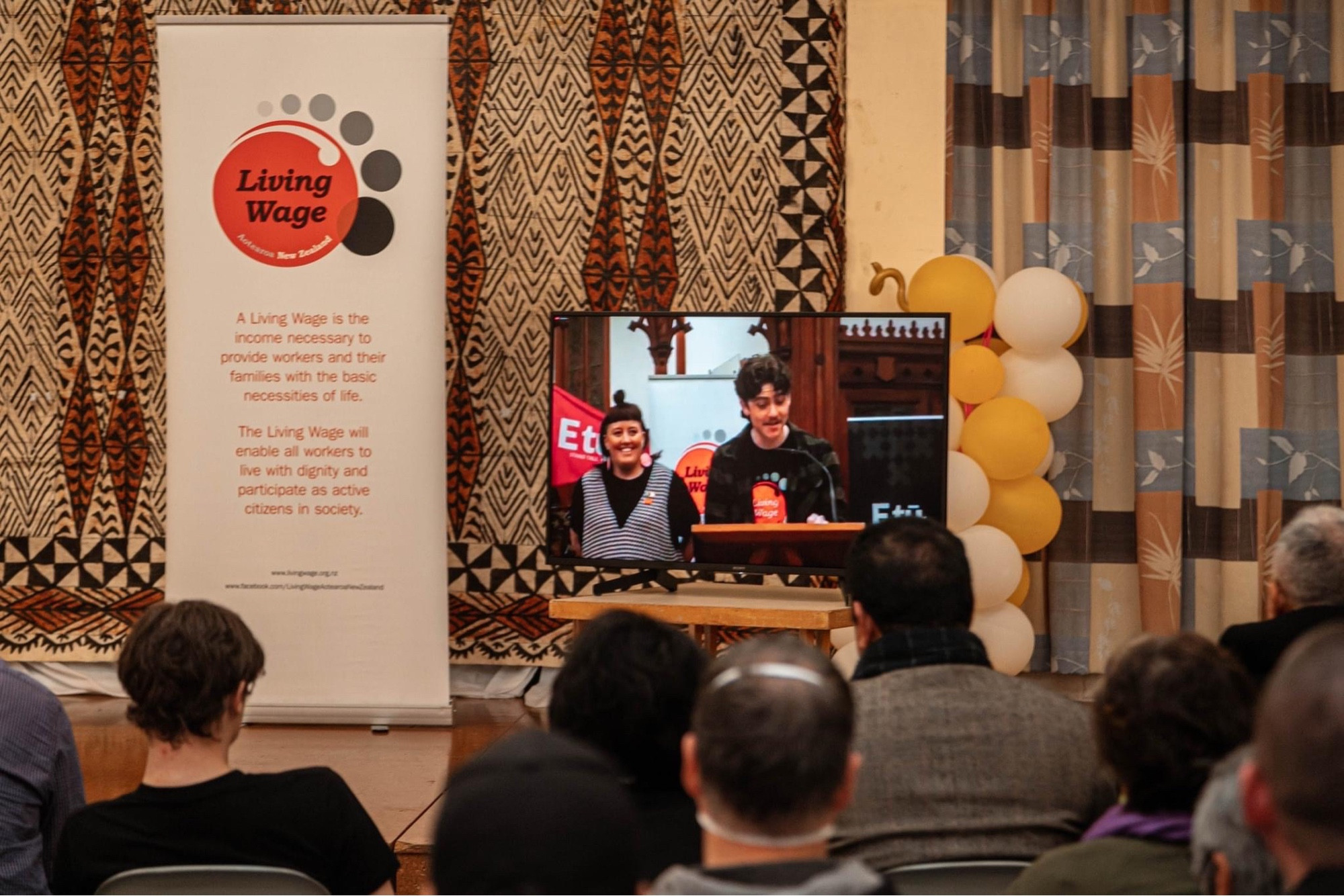 Photograph of Workers in the public sector in Wellington on screen in Christchurch addressing the crowd. There’s a Living Wage banner to the left of the screen in front of a tapa cloth. Two workers are on the screen, one is speaking. Yellow and white balloons are behind the screen to the right. In the foreground you can see the backs of some of the attendees heads.