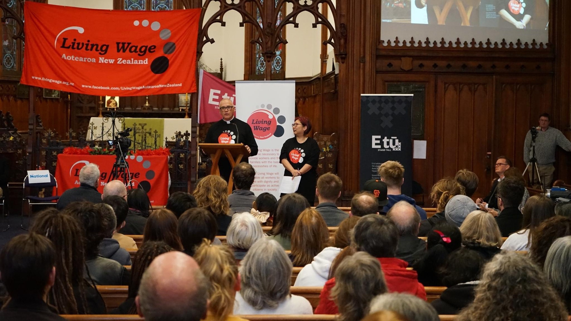 Photograph of attendees at the Wellington Living Wage Election Forum 2023 facing towards the speaker who is on the pulpit in St. Peters on Willis. Living Wage and Etuu banners are raised in the background.