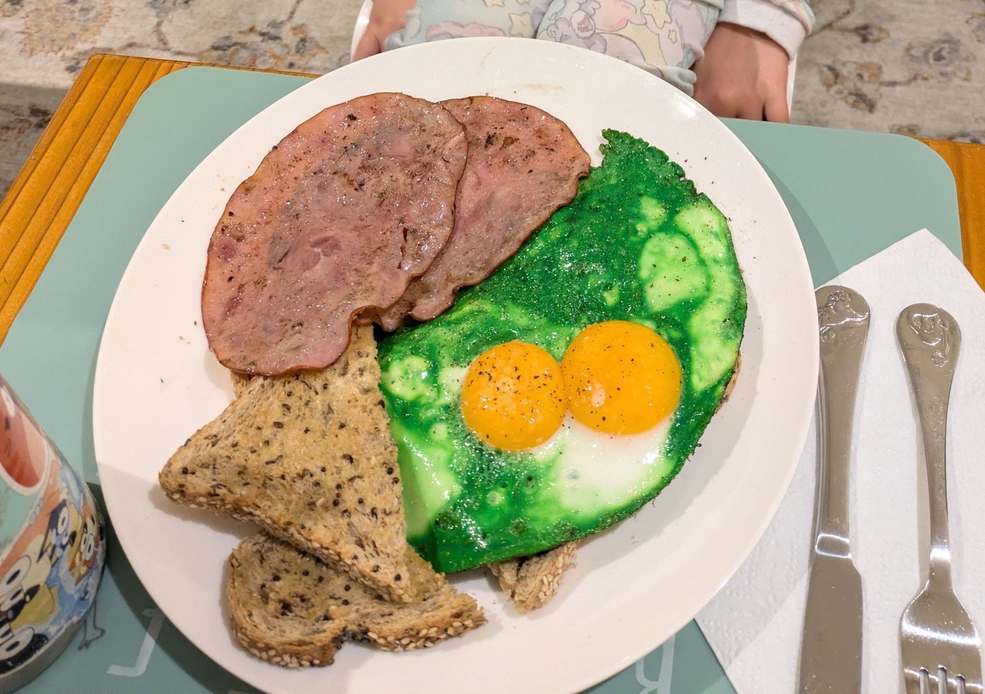 A plate of two fried eggs, died green, plus 2 slices of ham and multigrain toast. The plate is set upon an alphabet placemat with dinosaur cutlery and a water bottle covered in Bluey stickers.