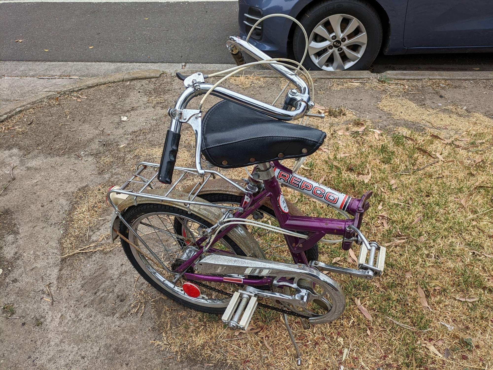 A vintage purple REPCO folding bike, all folded up. Despite its apparent age and the rust on the fenders, the hinges and other parts appear in pretty great shape.