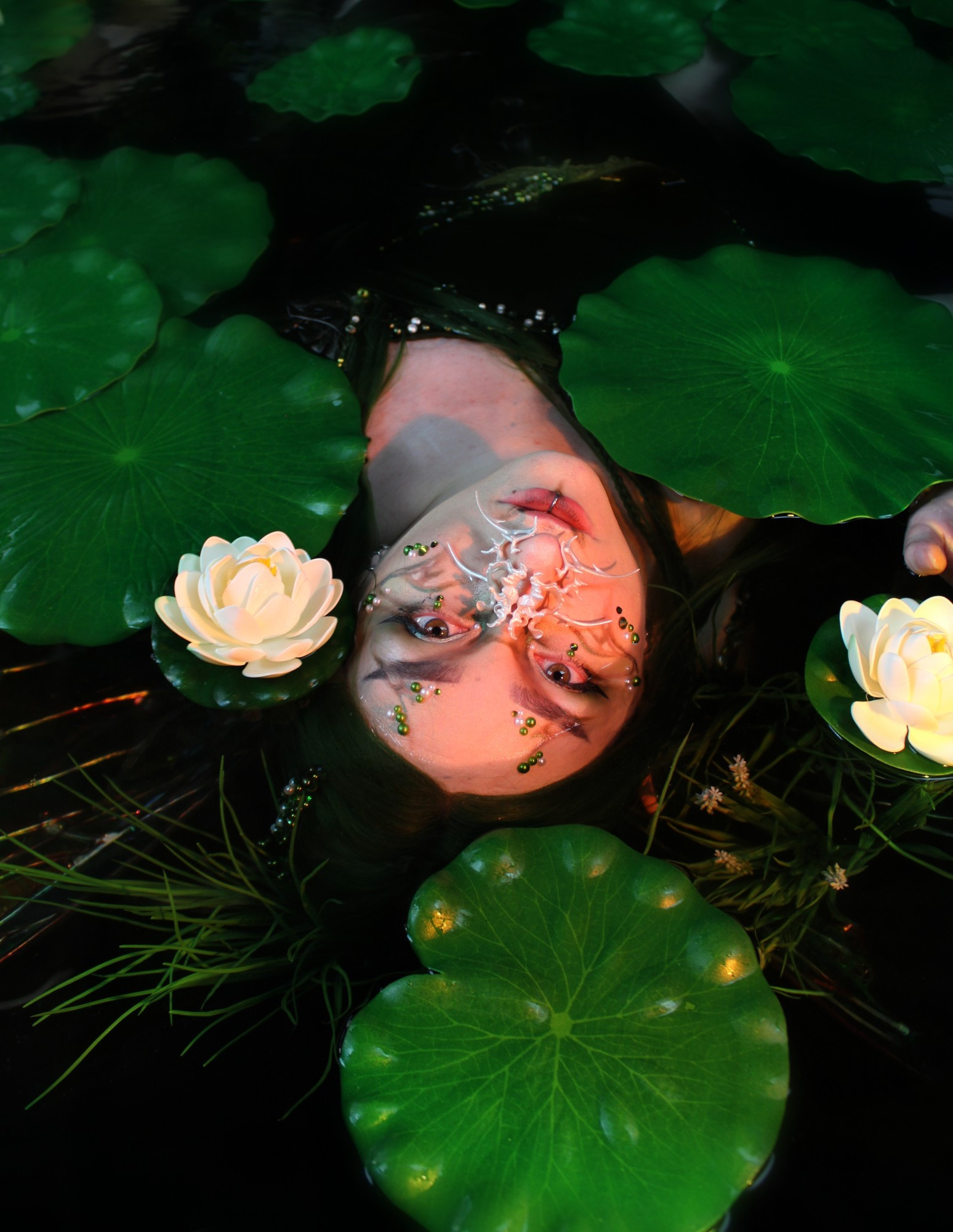 my face emerging from the water, surrounded by lily pads. I'm wearing an elaborate peal nose cuff and ornate makeup.