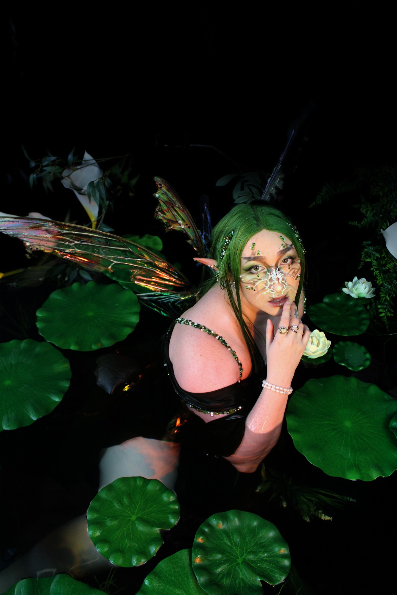 I'm sitting with my upper body emerging from the water, surrounded by lily pads. I have long green hair and holographic fairy wings.