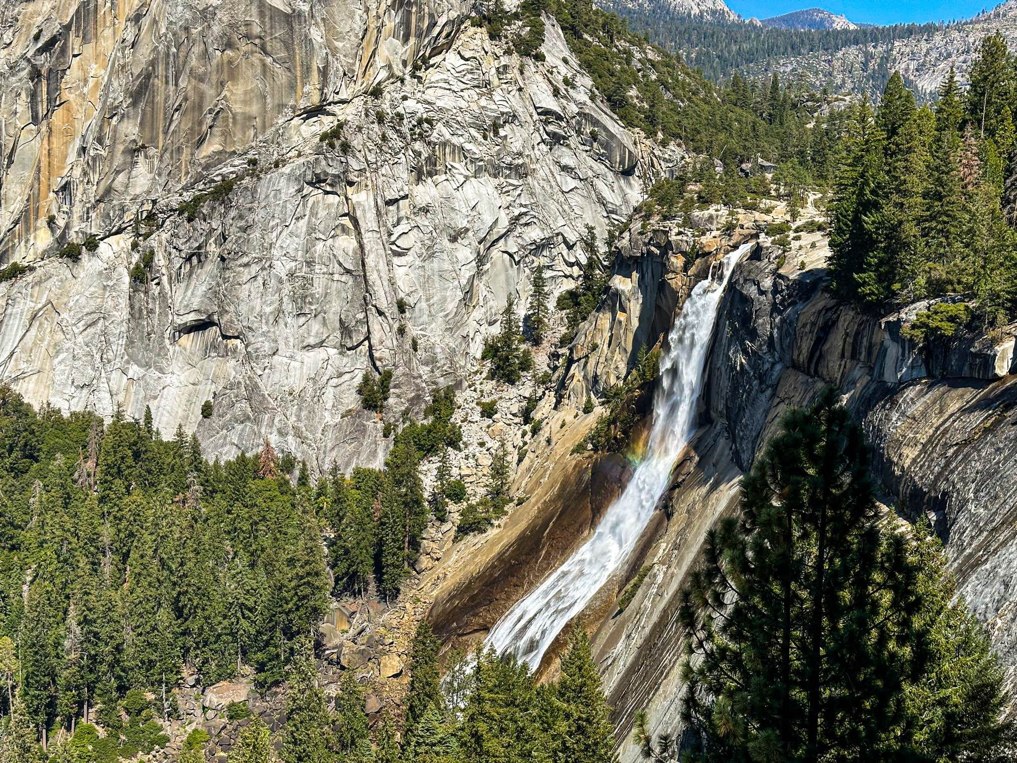 Nevada Fall seen from afar, with a tiny rainbow and mountains ans trees around it