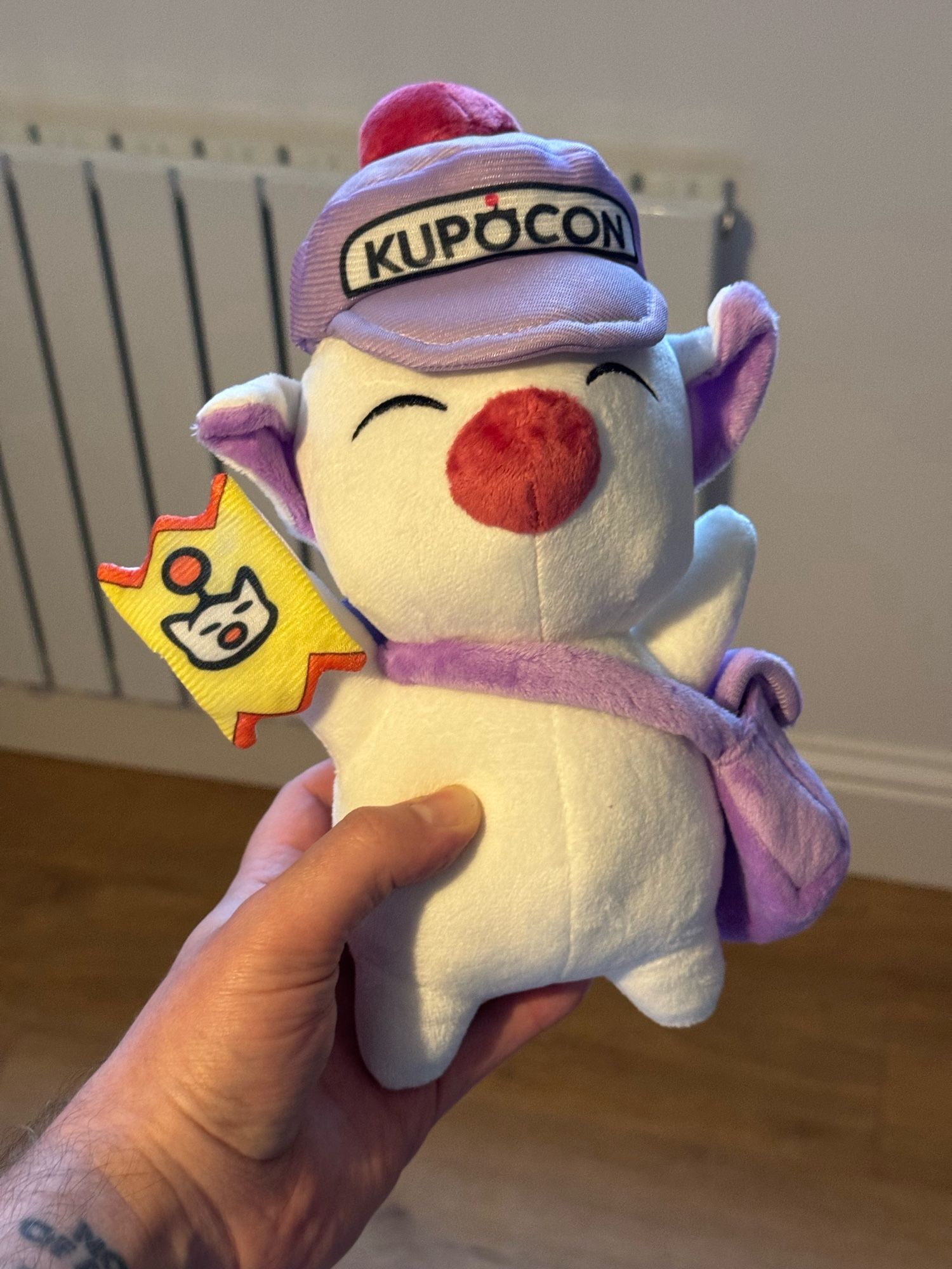 A photograph of me holding a Moogle plushie, a white fairy creature with a red nose from final fantasy. He’s wearing a purple cap reading “Kupocon”, and a purple knapsack. In his right hand is a yellow ticket with a moogle’s face on it. It’s all very cute