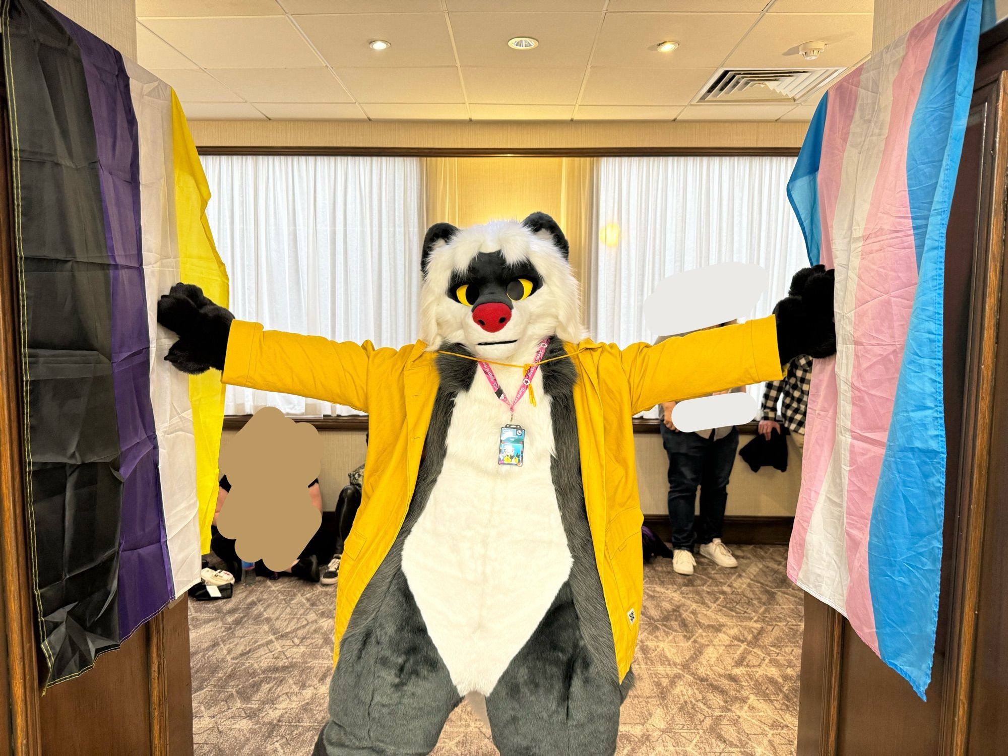 ConFuzzled’s mascot, Mel, standing proudly between a nonbinary flag and a trans flag