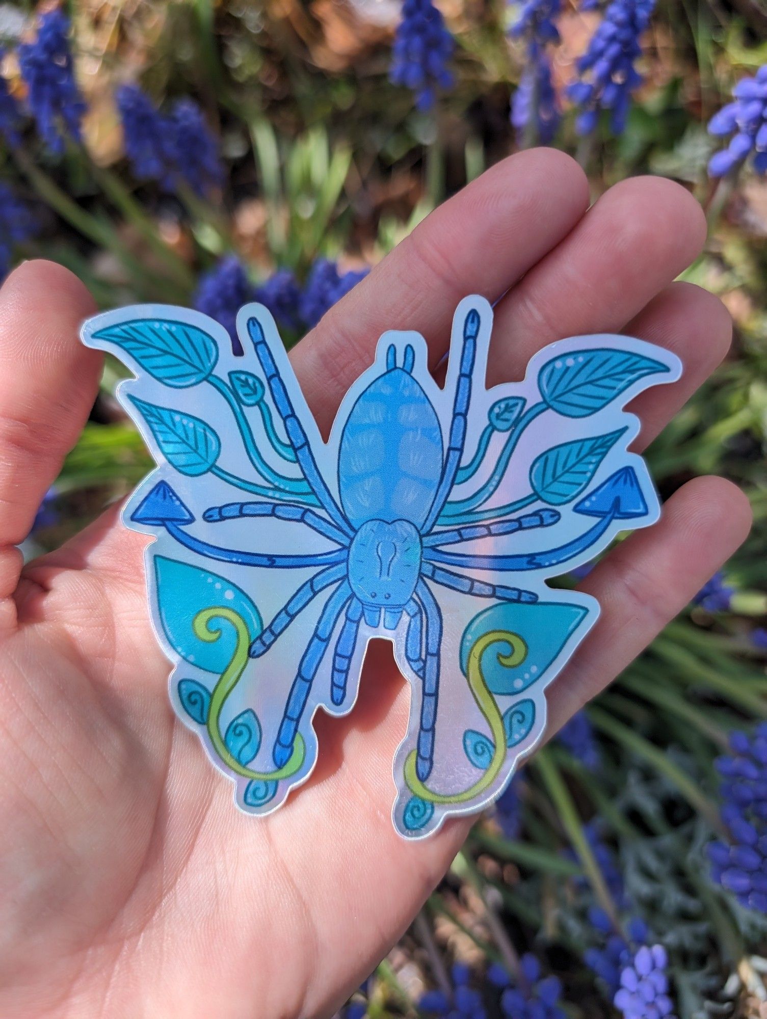 Holographic sticker consisting of a Caribena Versicolor Tarantula Sling surrounded by blue leaves and mushrooms.