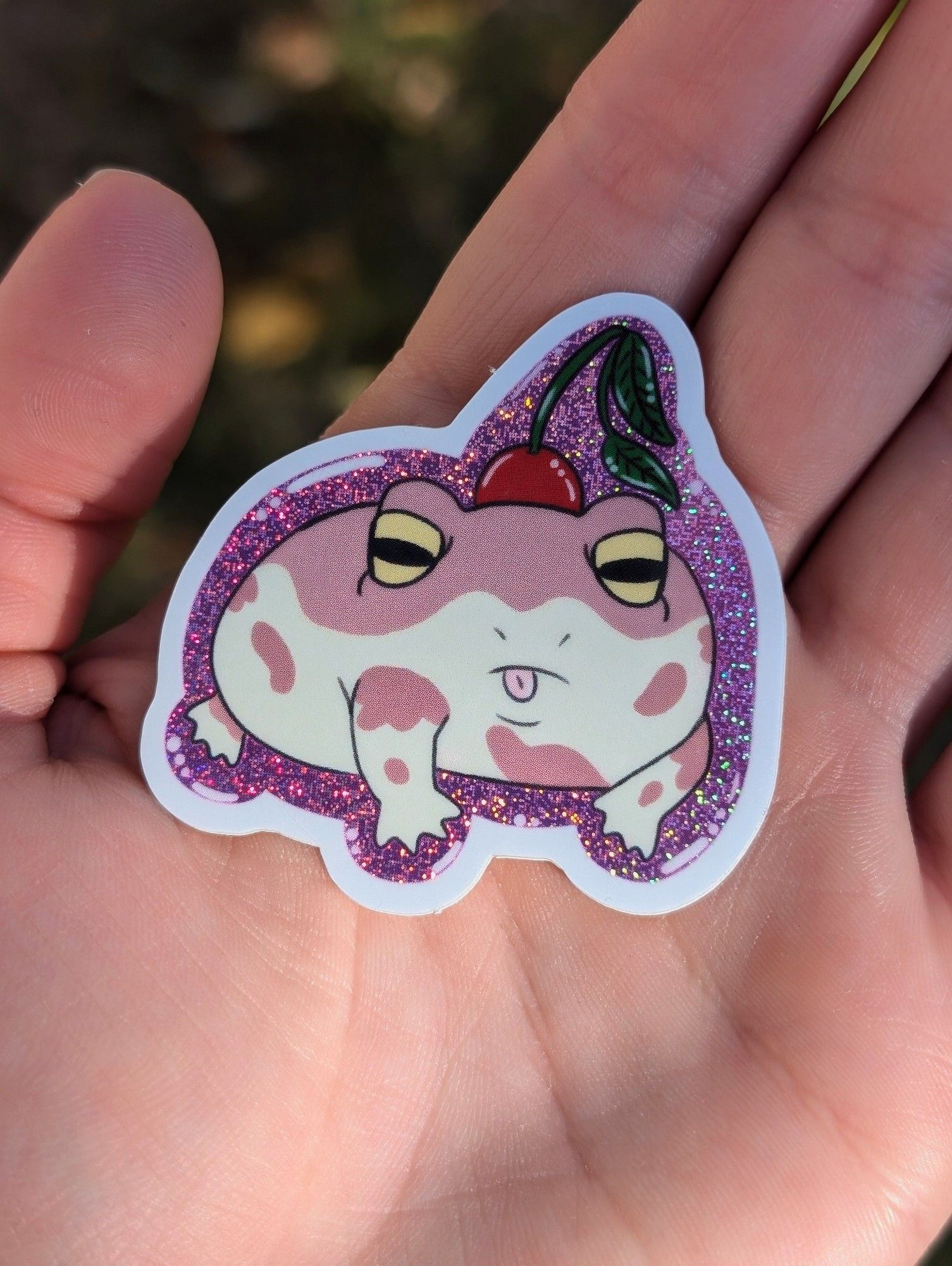 Glittery sticker featuring a Rain Frog sticking it's tongue out and wearing a cherry as a hat.