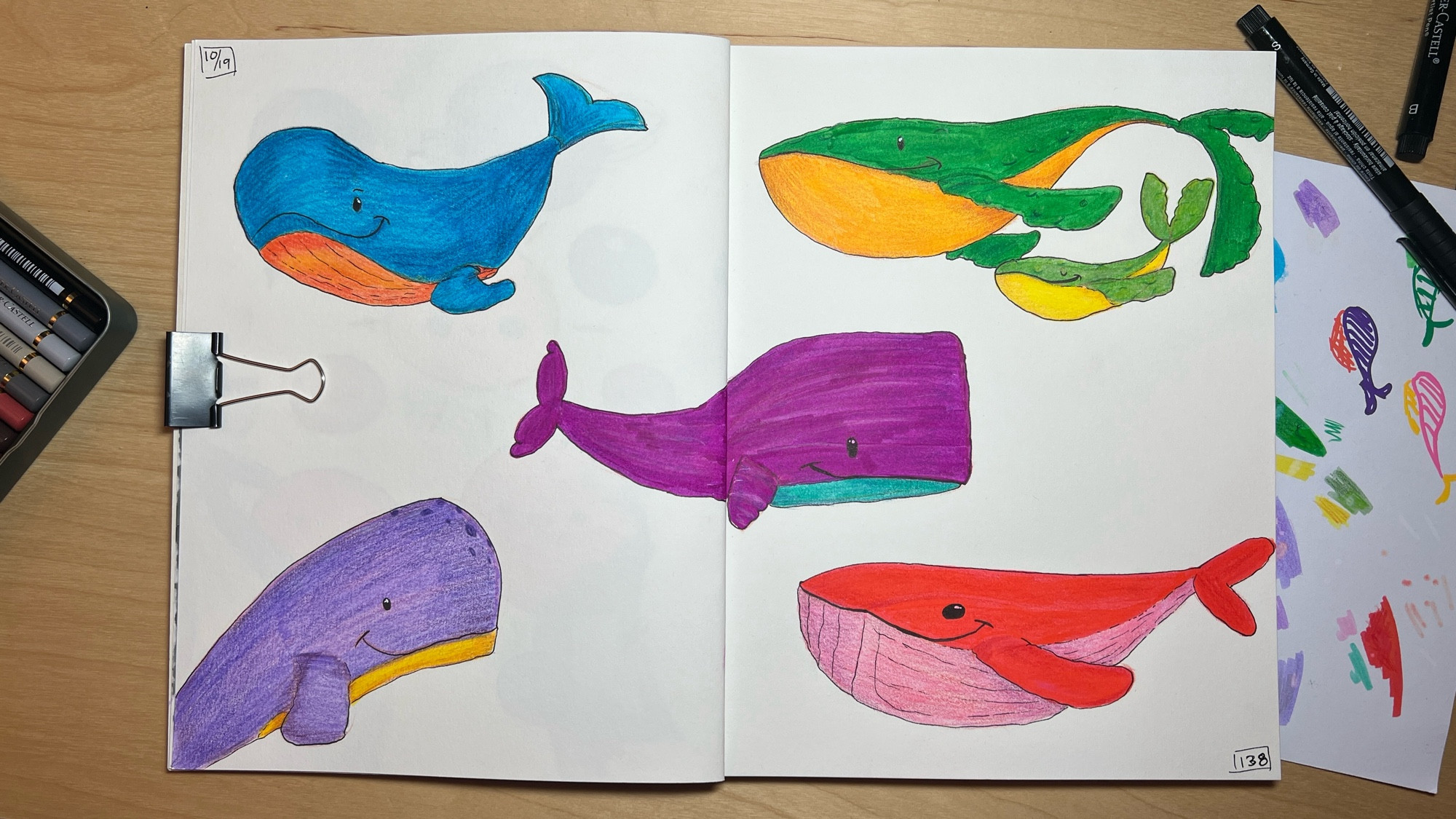 An open sketchbook on a table. The pages are filled with colorful sketches of whales in multiple bright colors like blue, orange, yellow, green, and purple.