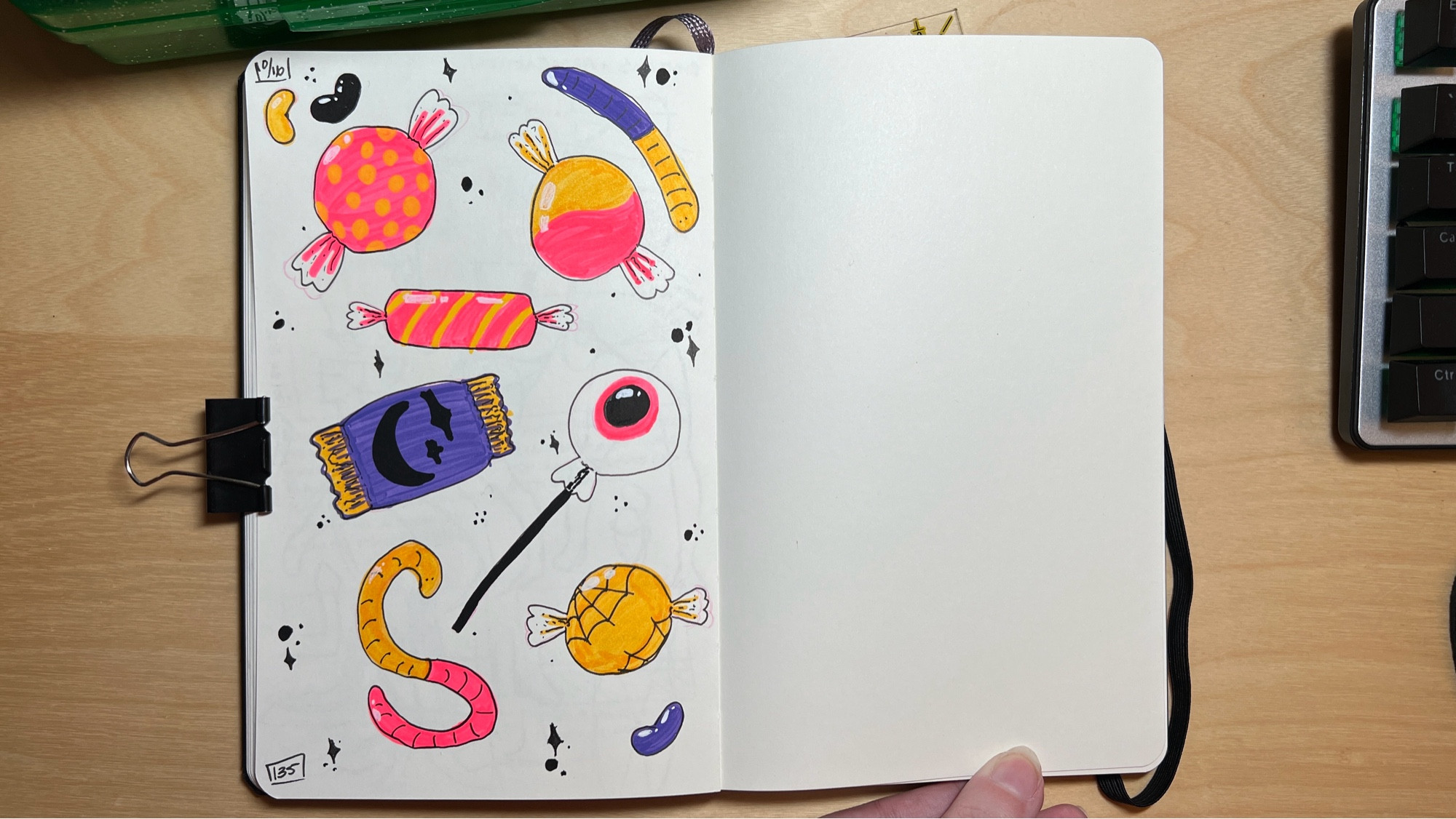 An open sketchbook on a table. The pages are filled with colorful sketches of candy: gummy worms, jelly beans, hard candy, and a packaged chocolate. Colors used are neon pink, yellow, purple, and black. Black dots and tiny stars fill in the blank spaces between the pieces of candy.