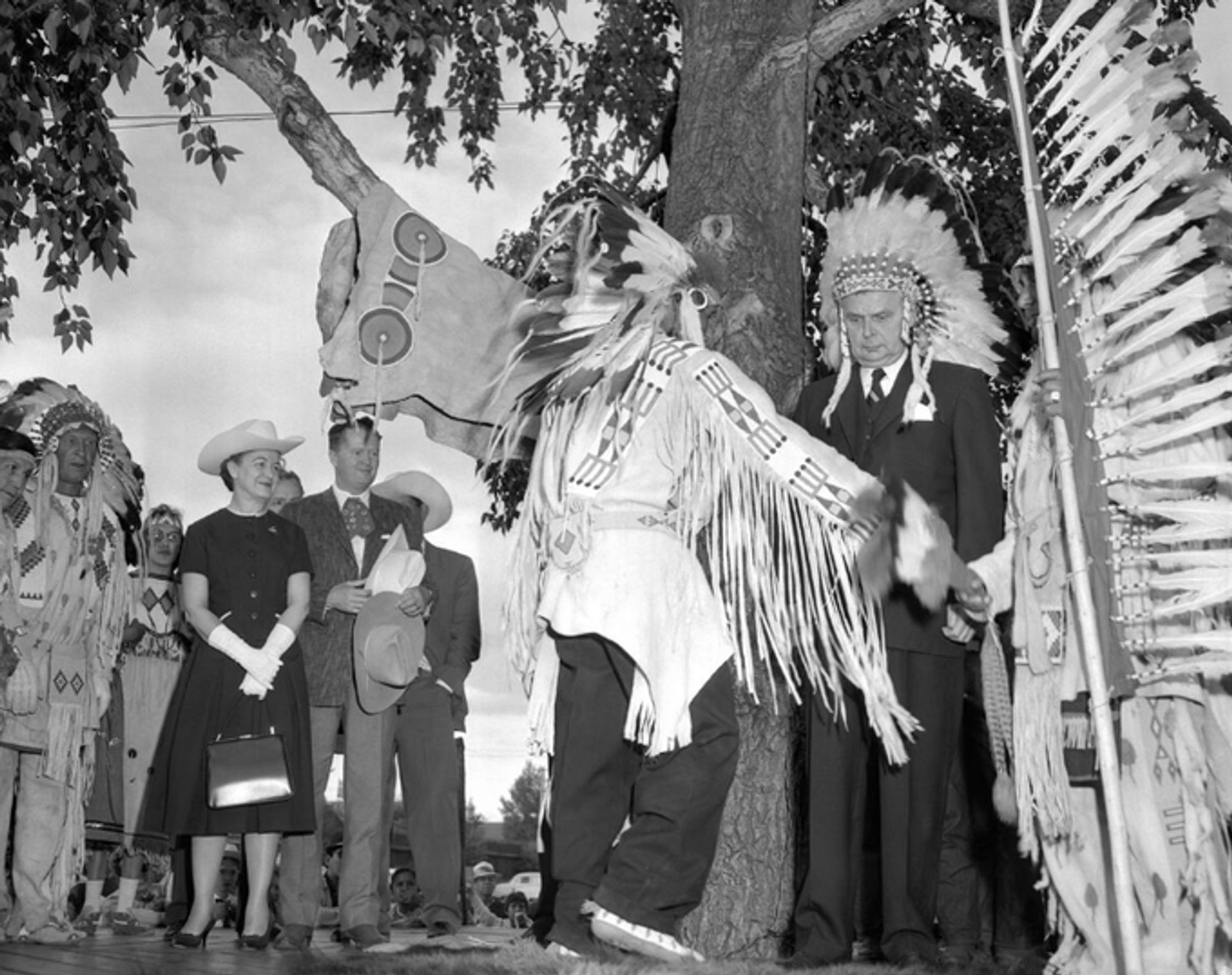 An older man in a suit wearing a headdress. A crowd is watching as another man in regalia performs.