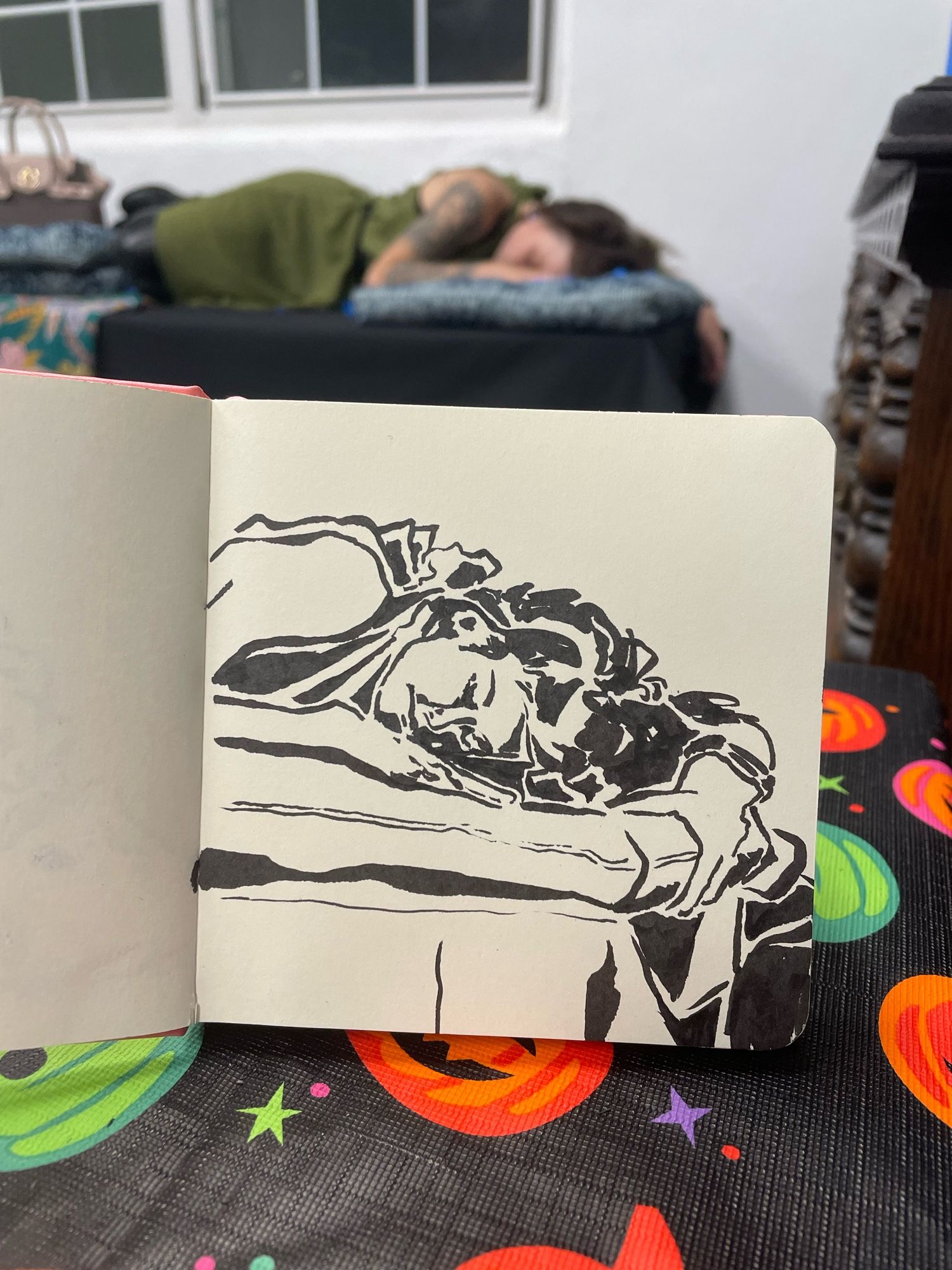 Ink sketch of a woman sleeping on a square pillow. She wears a watch and clutches the pillow with her left hand. Her hair falls over her arm. Her green dress ties at the back of her neck. The model is asleep, posing behind the drawing.